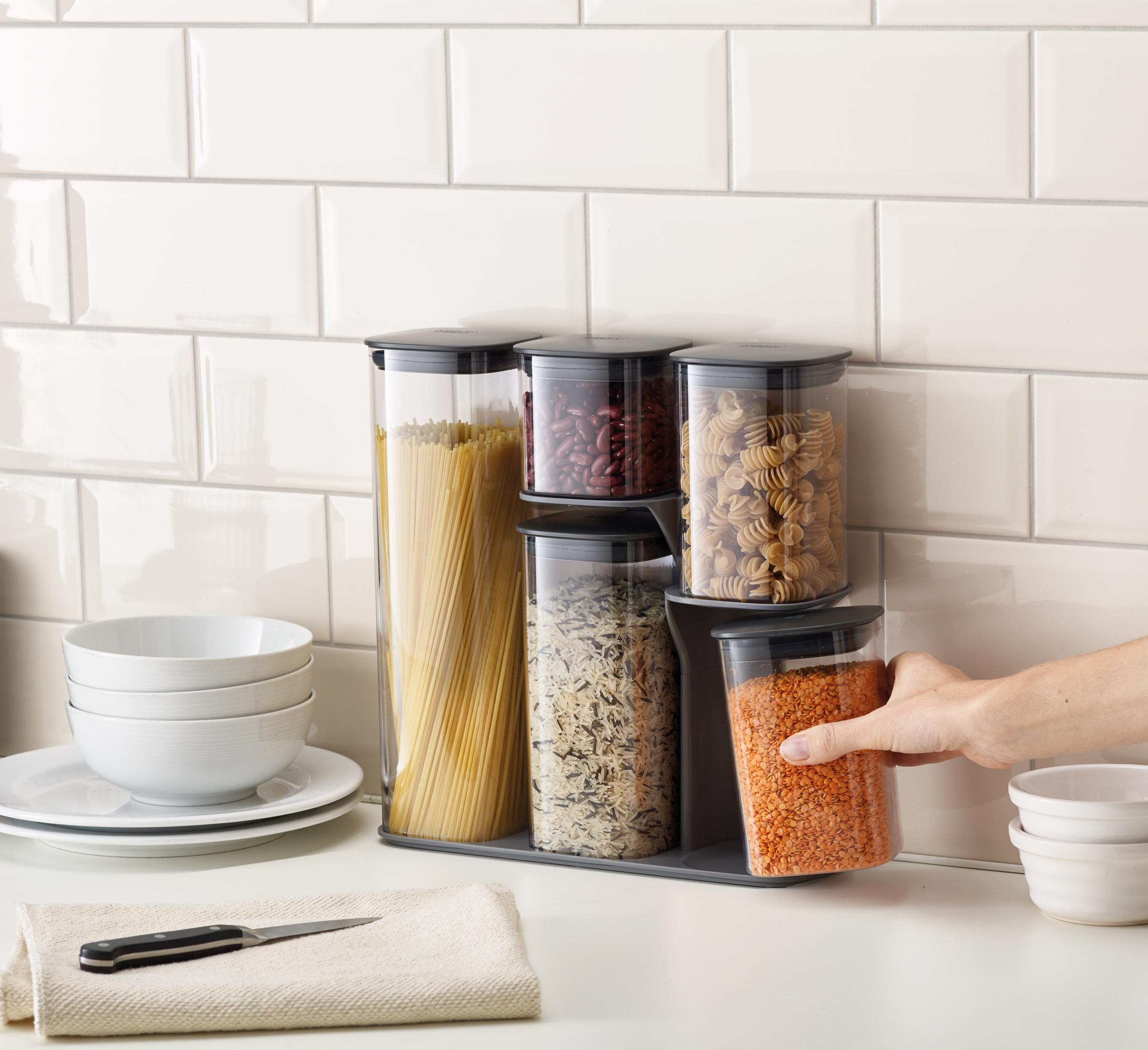 Recipientes de almacenamiento transparentes llenos de comida, dispuestos contra un protector contra salpicaduras de azulejos blancos.