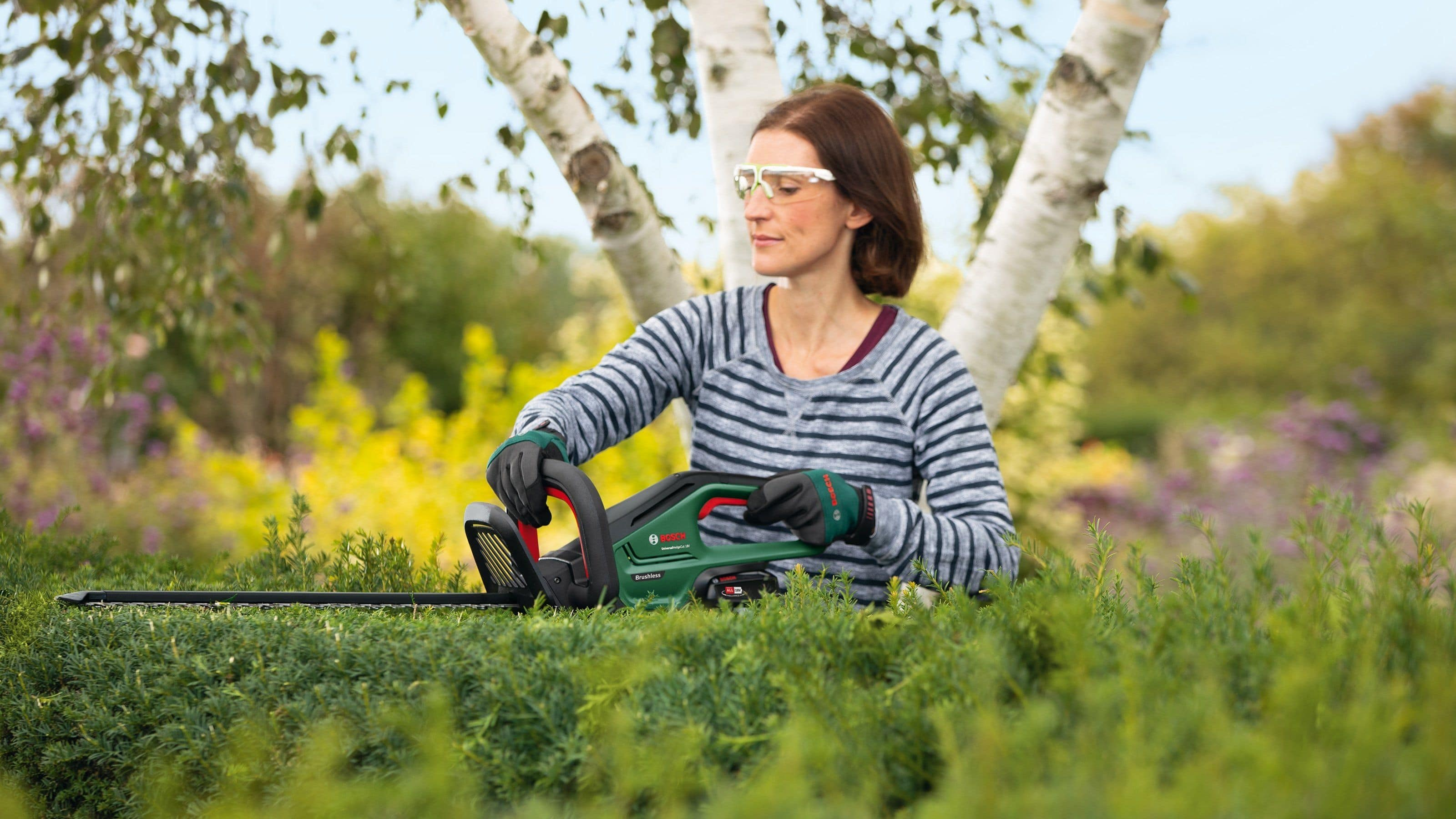 Eine Frau mit Schutzbrille benutzt eine grüne Heckenschere an einer grünen Hecke vor einem Baum.