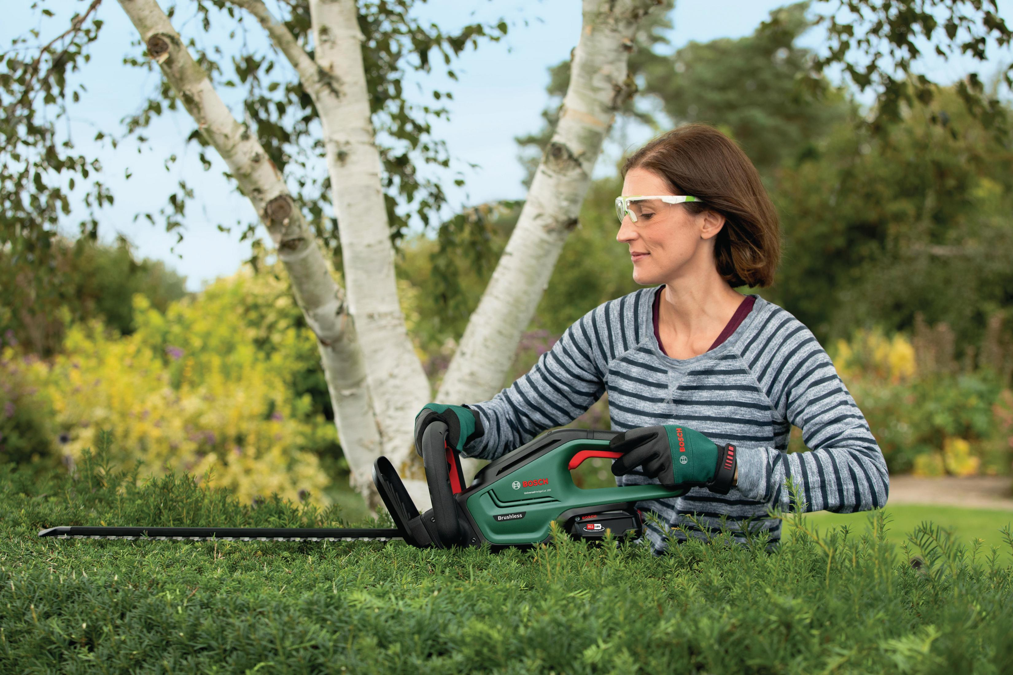 Eine Frau benutzt eine grüne Heckenschere an einer grünen Hecke vor einem Baum, während sie eine Schutzbrille trägt.