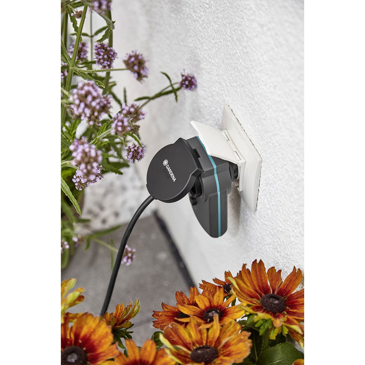 Adattatore di alimentazione intelligente Gardena collegato a una presa a muro con fiori intorno.