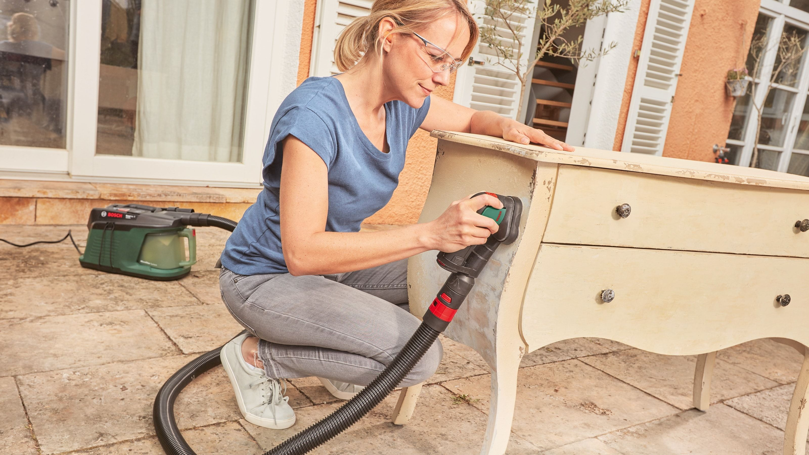 Eine Frau schleift mit einem Elektrowerkzeug eine beige Kommode ab, trägt eine Schutzbrille und Jeans. Draußen auf einer Steinterrasse.