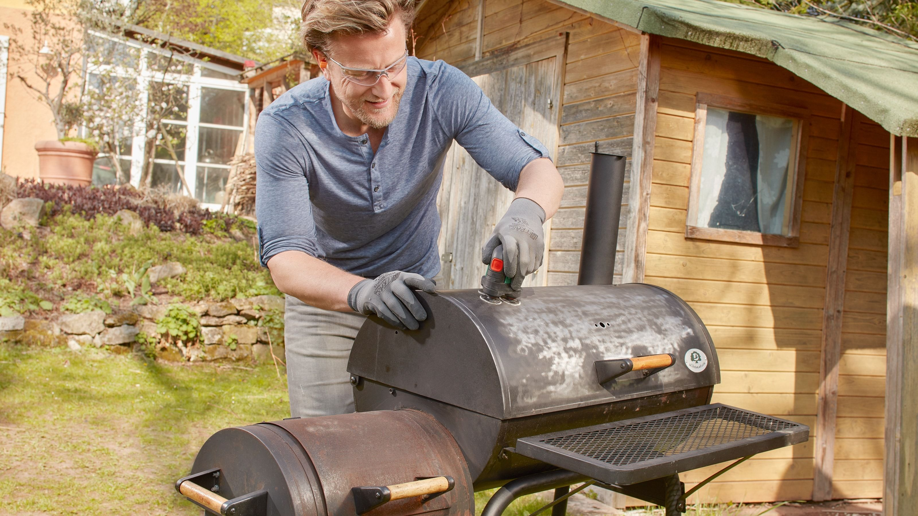 Ein Mann schleift im Freien mit einem Elektrowerkzeug einen schwarzen Grill ab, trägt dabei eine Schutzbrille und Handschuhe neben einem Holzschuppen.