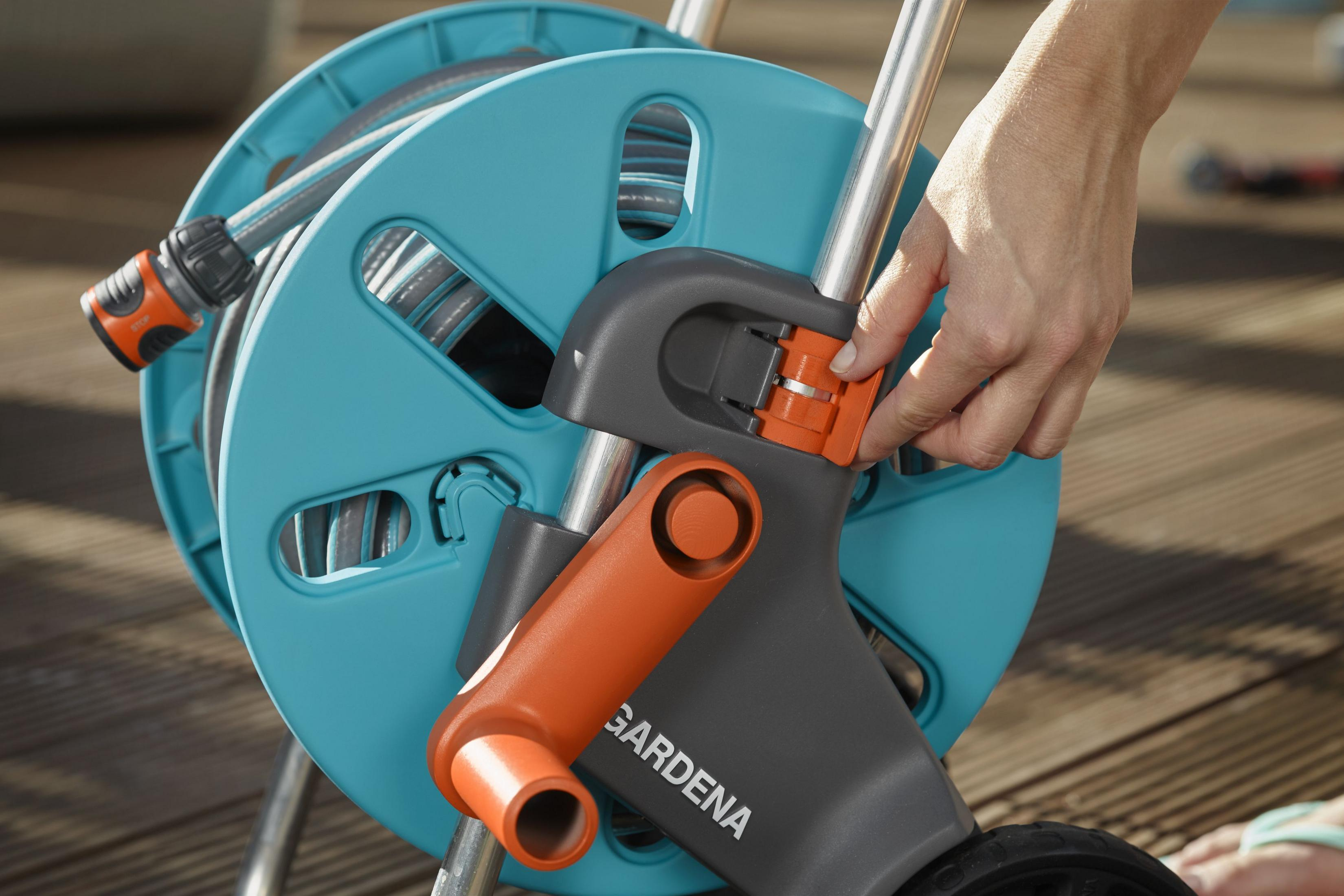 Nahaufnahme einer Hand, die einen Gardena Schlauchaufroller mit blauer Trommel und orangefarbenem Griff einstellt.