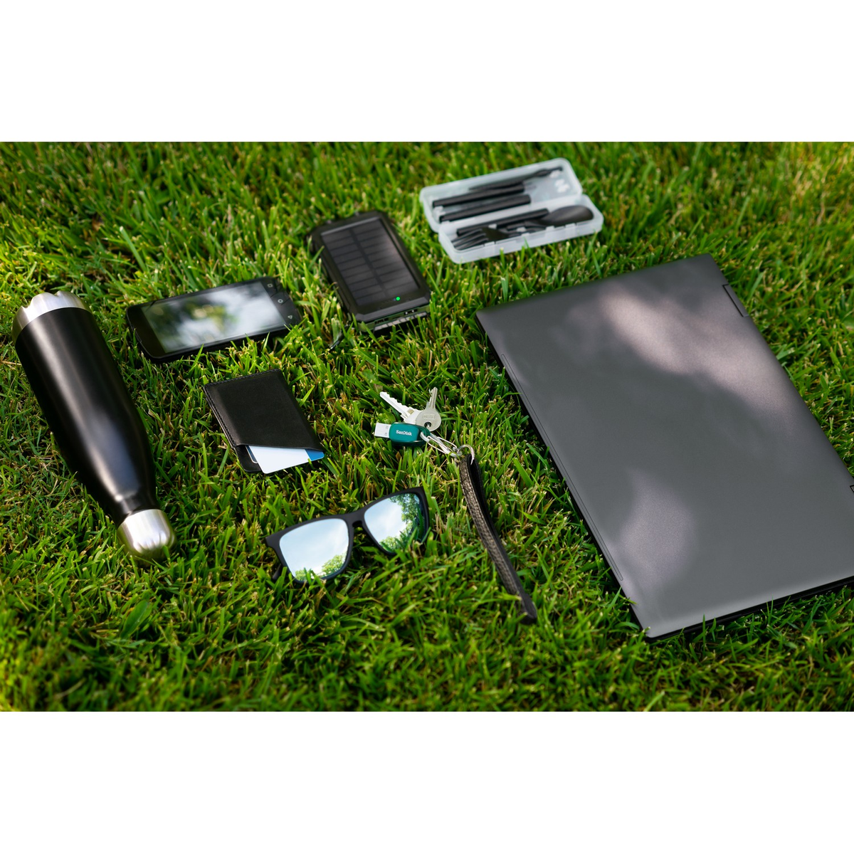 Scène d'extérieur : ordinateur portable, téléphone, bouteille, portefeuille, clés, lunettes, chargeur solaire, jeu de tournevis sur herbe verte.