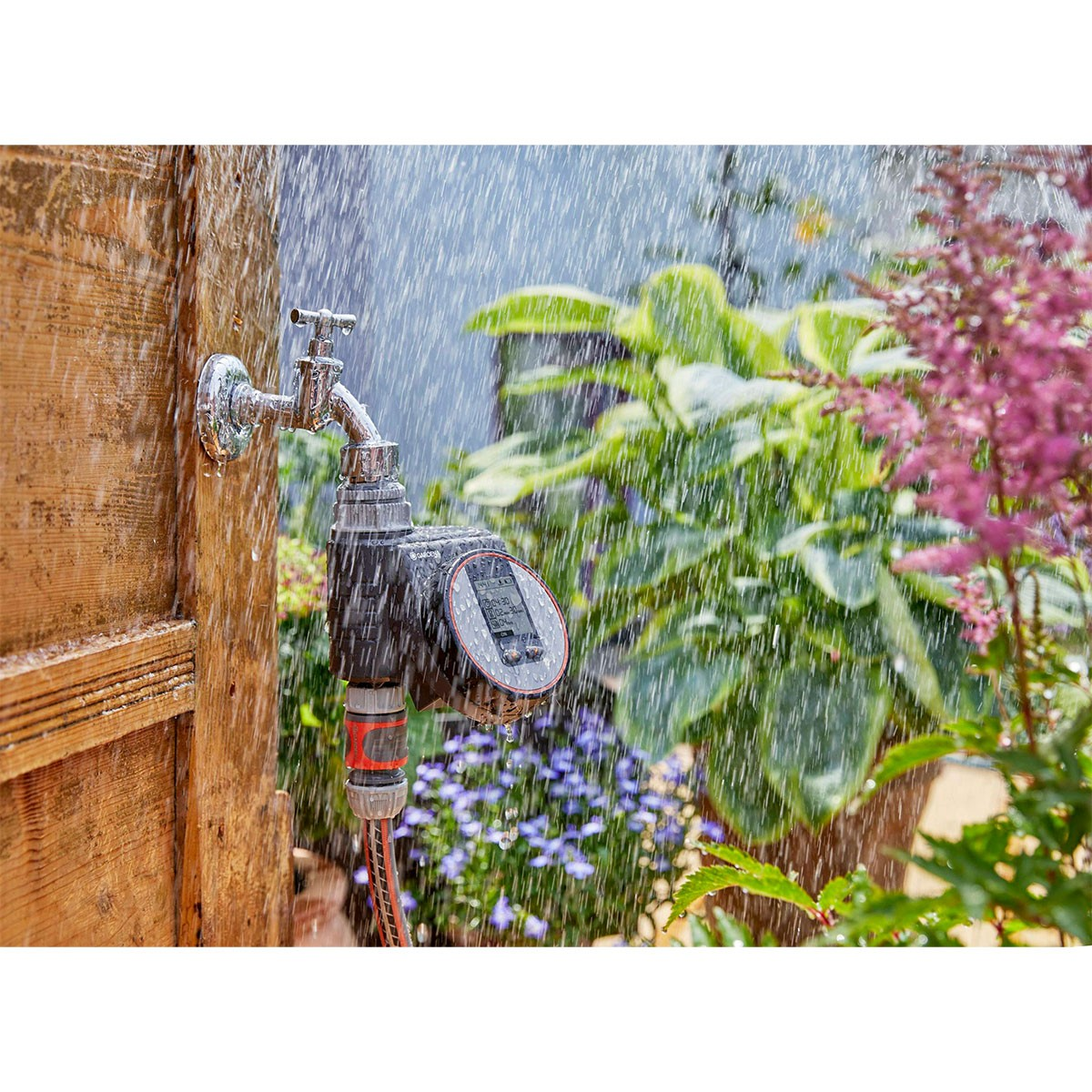 Gardena-Bewässerungstimer an einem Wasserhahn mit Wassersprühnebel. Grüne Pflanzen und Blumen im Hintergrund.