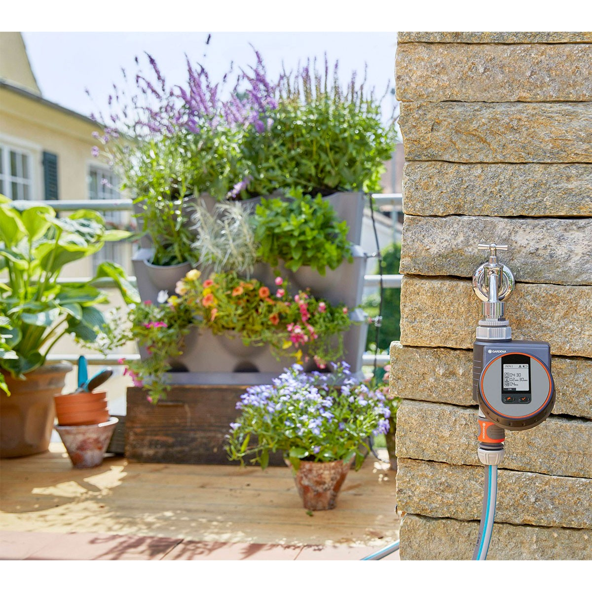 Gardena-Bewässerungstimer an einem Wasserhahn, mit Pflanzen und Blumen im Hintergrund auf einem Balkon.