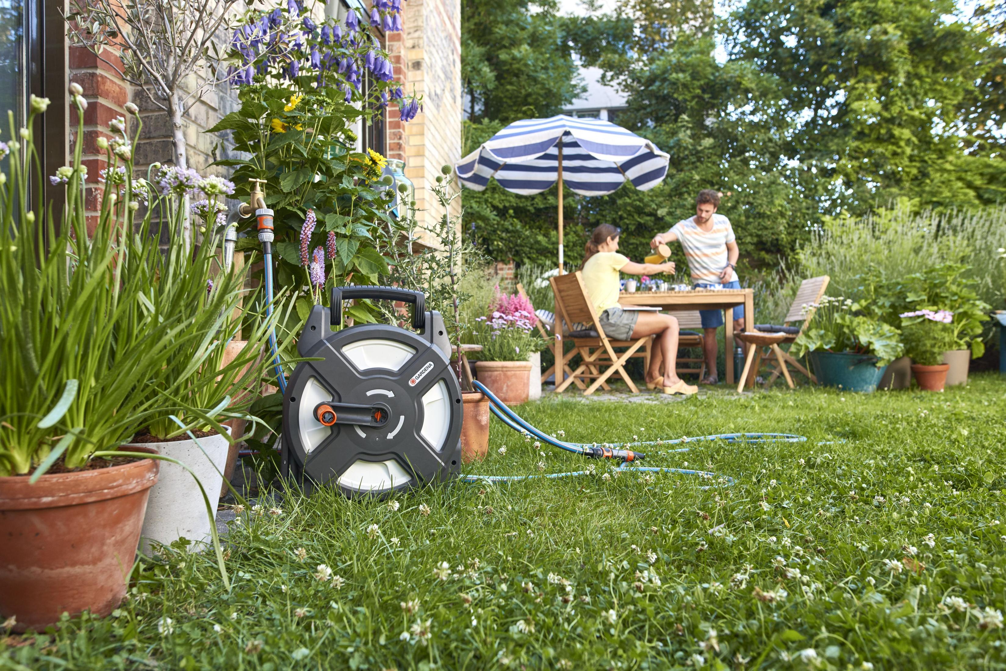 Eine Gardena Schlauchbox steht auf einem Rasen in der Nähe eines Gartentisches, an dem unter einem Sonnenschirm gegessen wird.