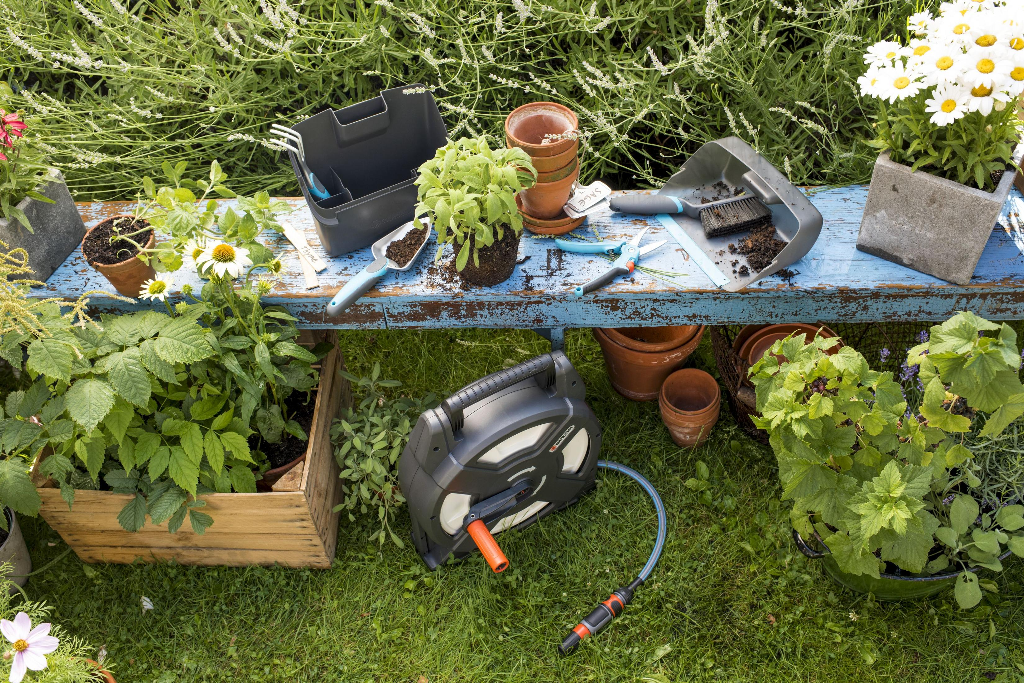 Gartenszene mit einer Schlauchhaspel, Pflanzen und Gartengeräten auf einer blauen Bank vor einem grünen Rasen.
