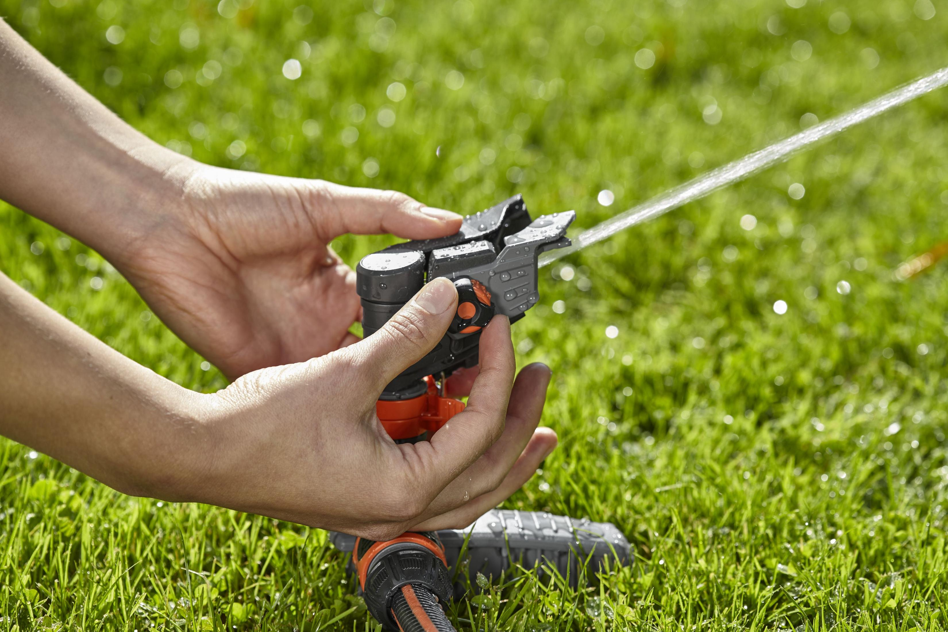 Hände justieren einen orangefarbenen und grauen Sprinkler auf grünem Gras, der Wasser versprüht.