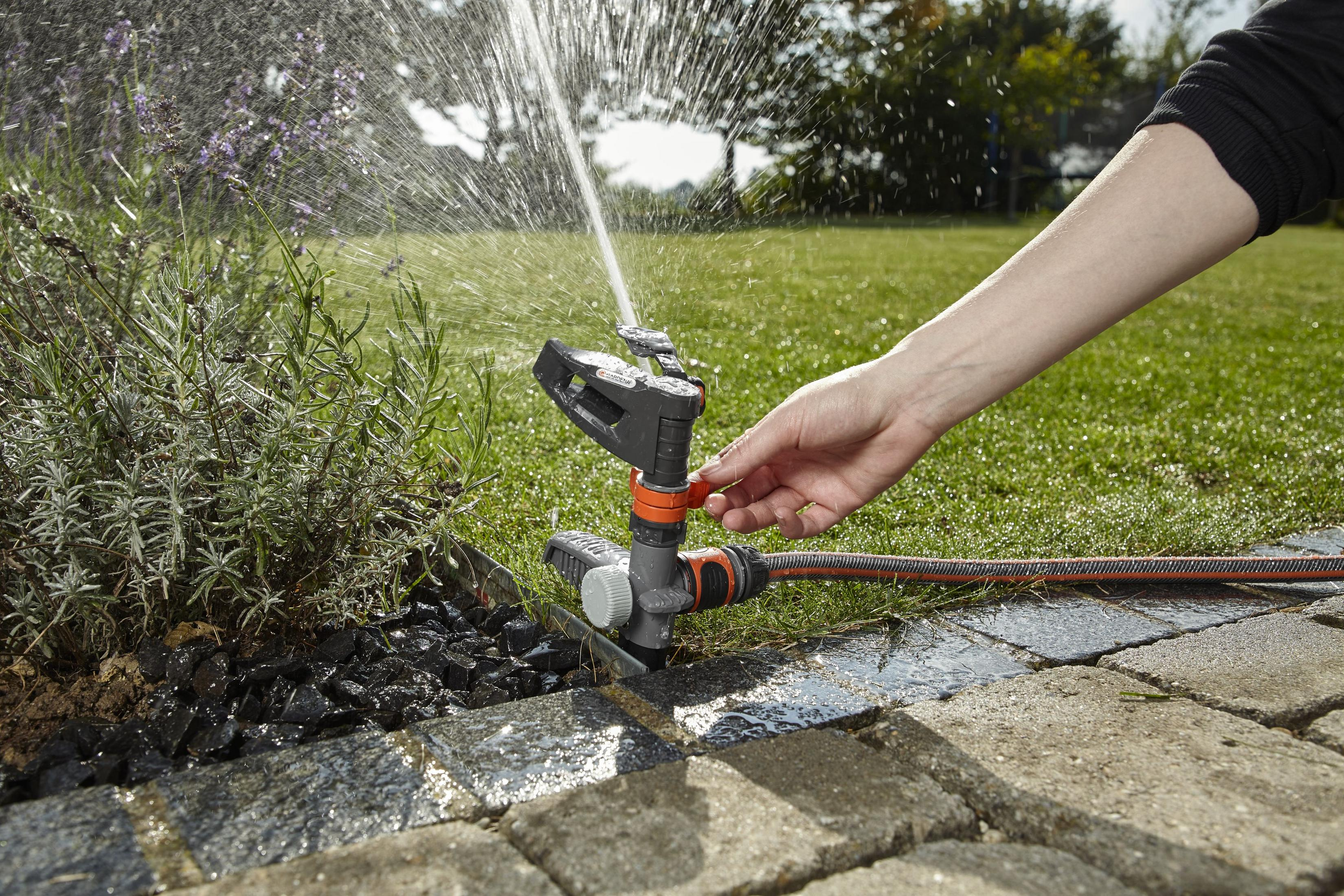 Ein Sprinkler bewässert einen Garten mit Grün und Steinplatten und versprüht Wasser.