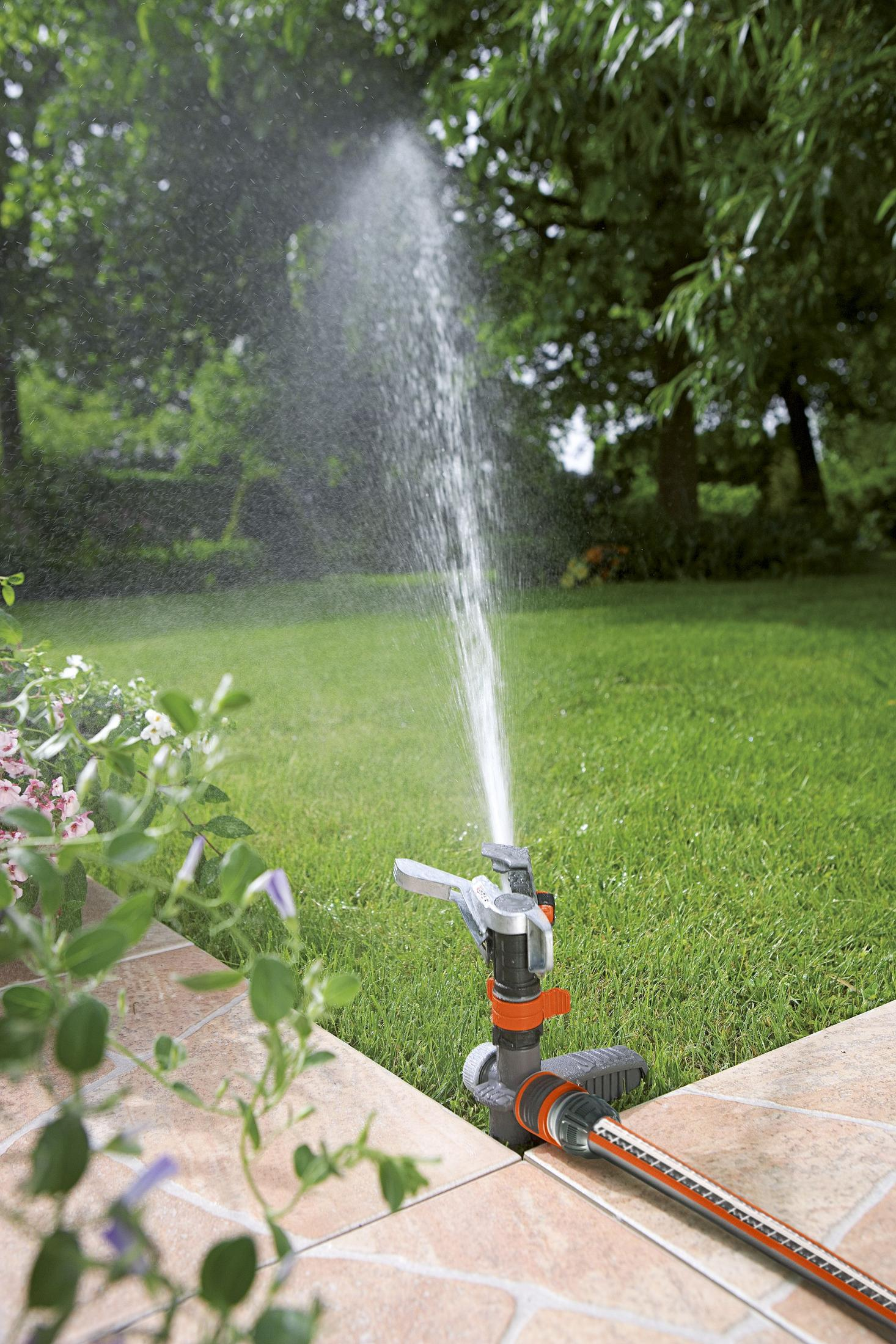Ein Sprinkler bewässert einen Garten mit Grün und Steinplatten und versprüht Wasser.