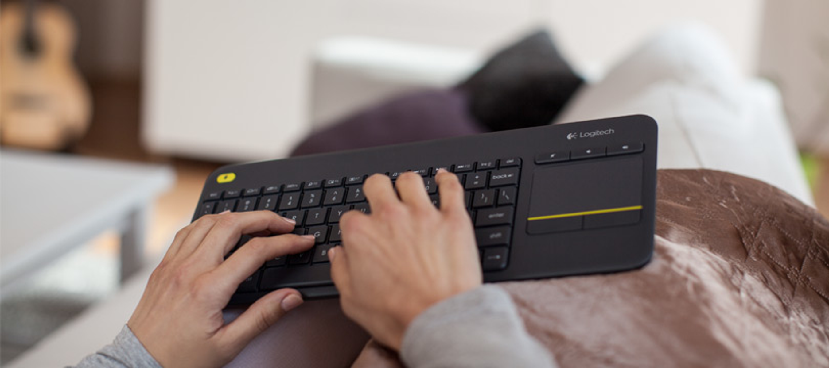 Hände, die auf einer schwarzen Logitech-Tastatur mit Touchpad tippen, ruhen auf dem Schoß einer Person. Der Hintergrund ist unscharf.