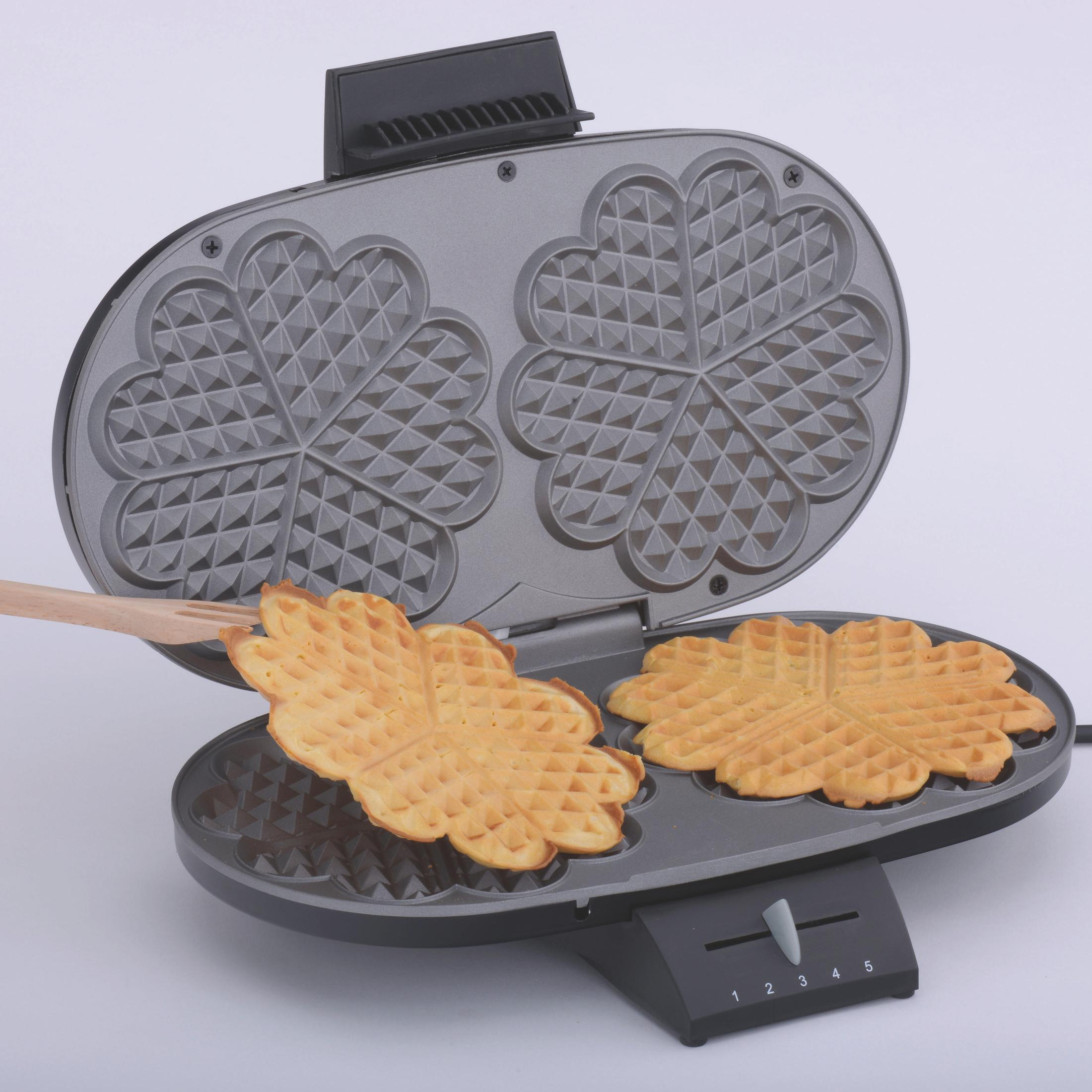 Otwarta gofrownica z dwoma goframi w kształcie serca wyjmowanymi za pomocą drewnianego narzędzia.