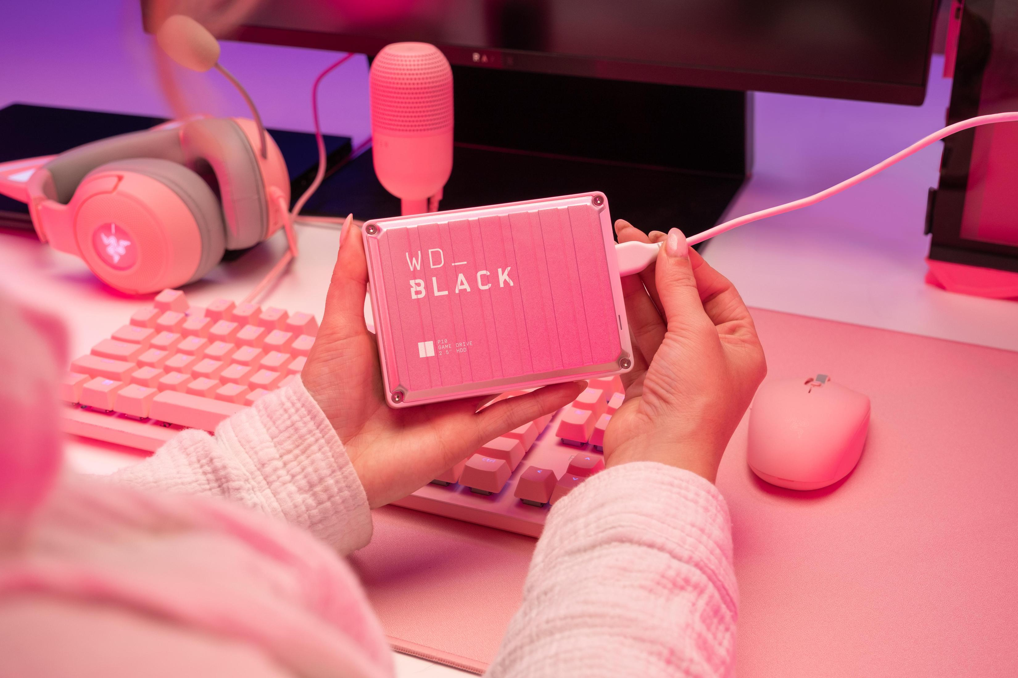 Un disco duro WD Black P10 rosa es sostenido por manos cerca de una configuración de juego rosa.