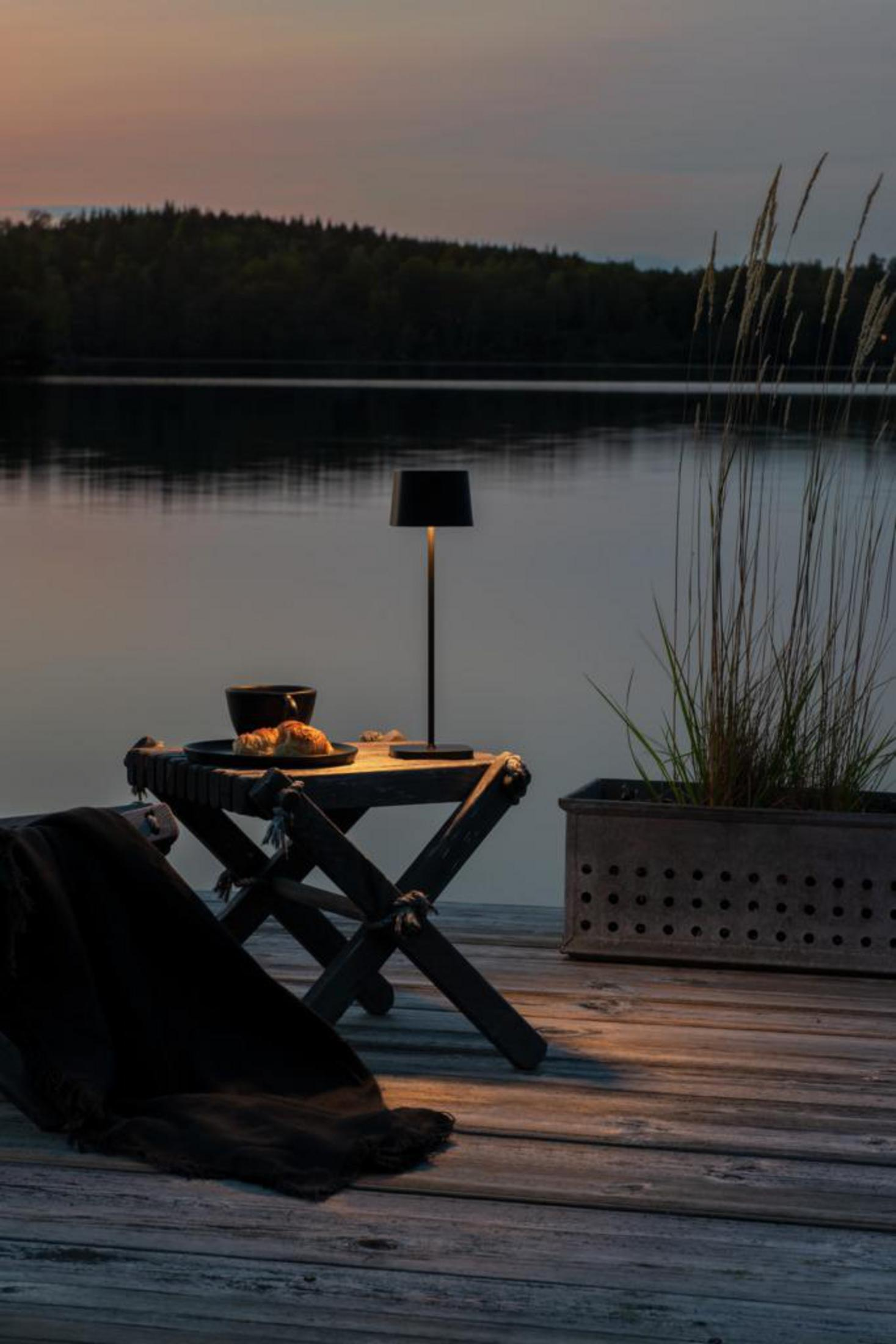 Außenszene: Lampe auf dem Tisch am See in der Dämmerung, mit Stuhl, Decke und Pflanze, die ein ruhiges Ambiente schafft.