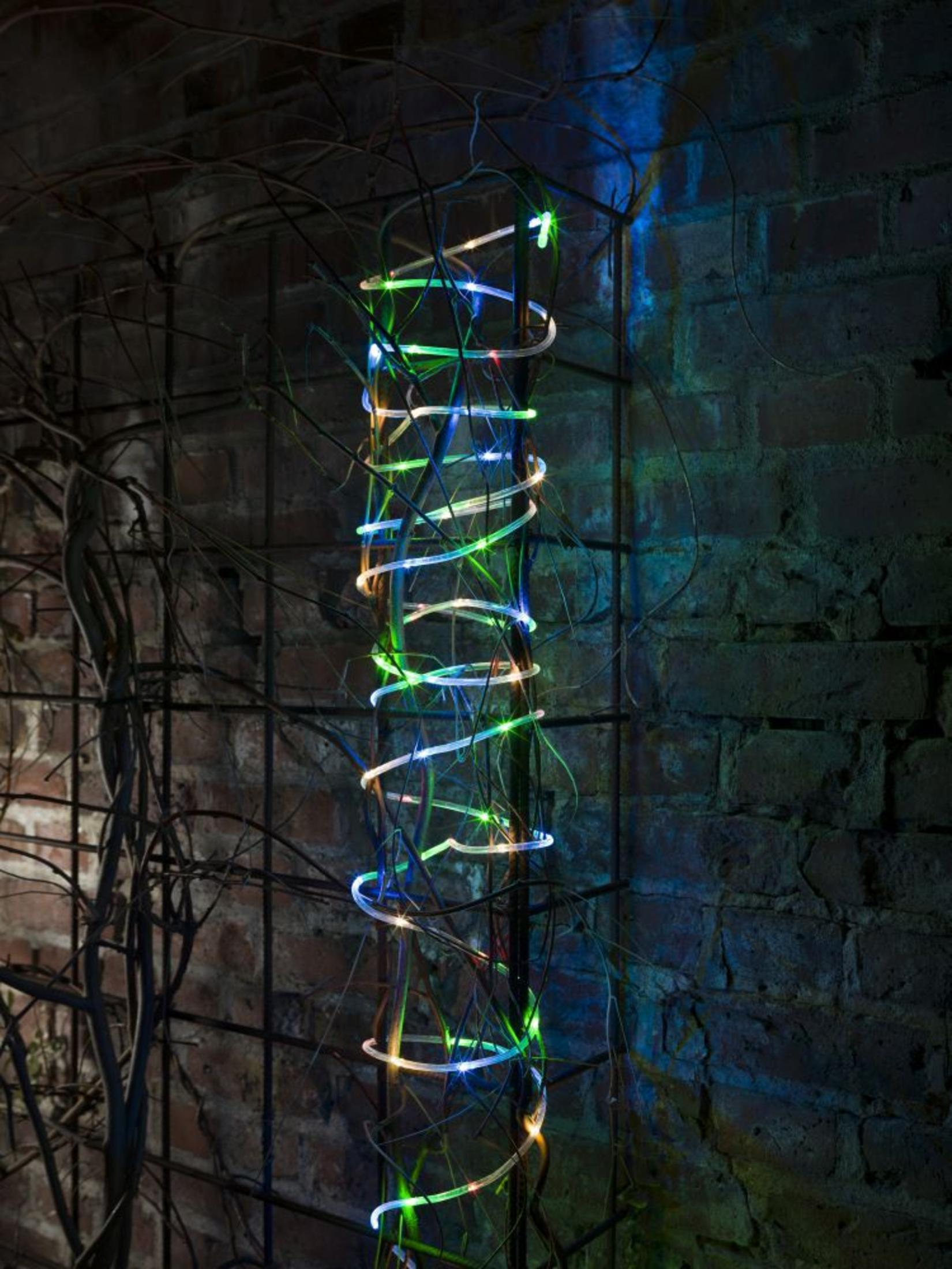 Eine Spirale aus LED-Lichtern windet sich um eine dunkle Metallstruktur vor einer Ziegelmauer.