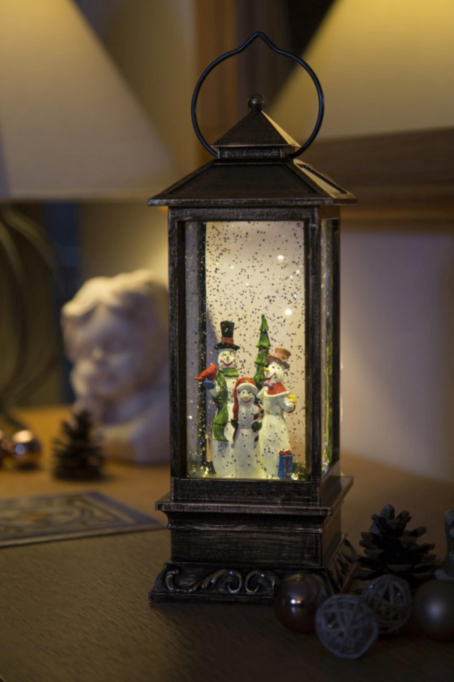 Eine dekorative Laterne mit Schneemännern im Inneren, auf einem Holztisch mit Ornamenten und einer Lampe im Hintergrund.