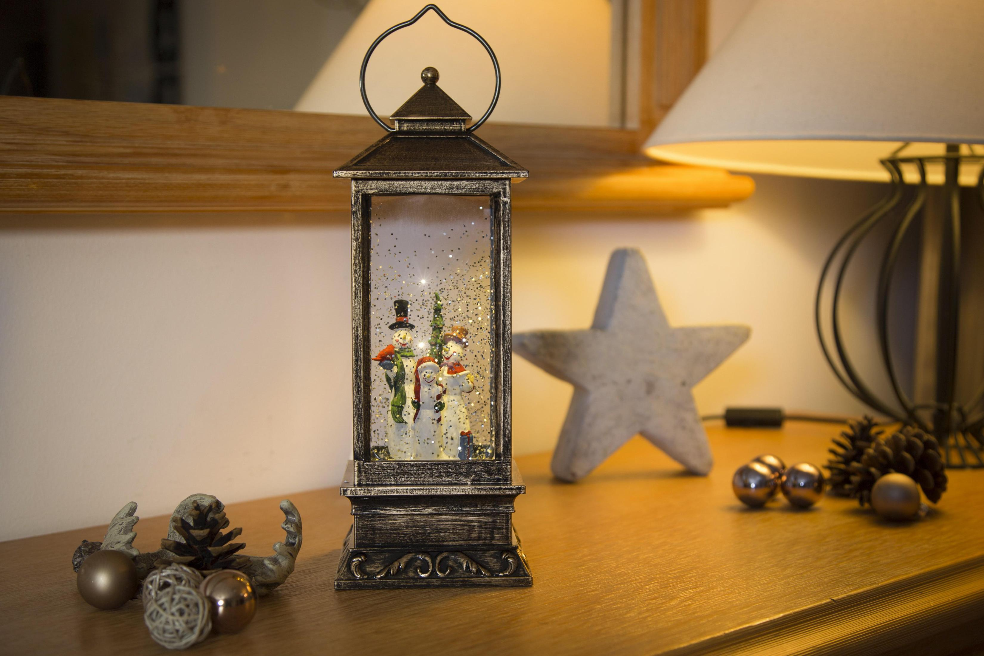 Eine dekorative Laterne mit Schneemännern im Inneren, auf einem Holztisch mit Ornamenten und einer Lampe im Hintergrund.