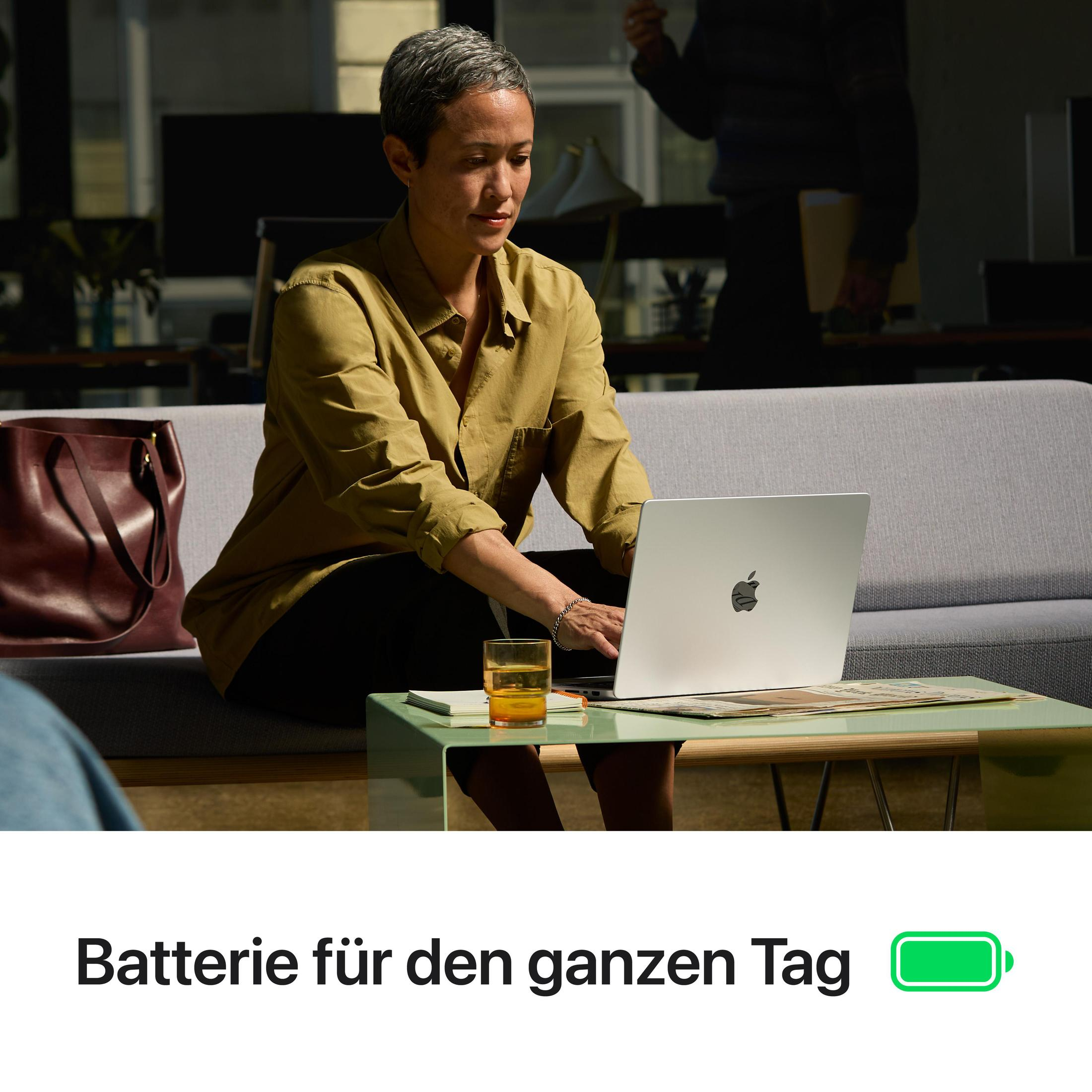 Eine Frau mit kurzen grauen Haaren arbeitet an einem silbernen Laptop, während sie auf einer grauen Couch sitzt.