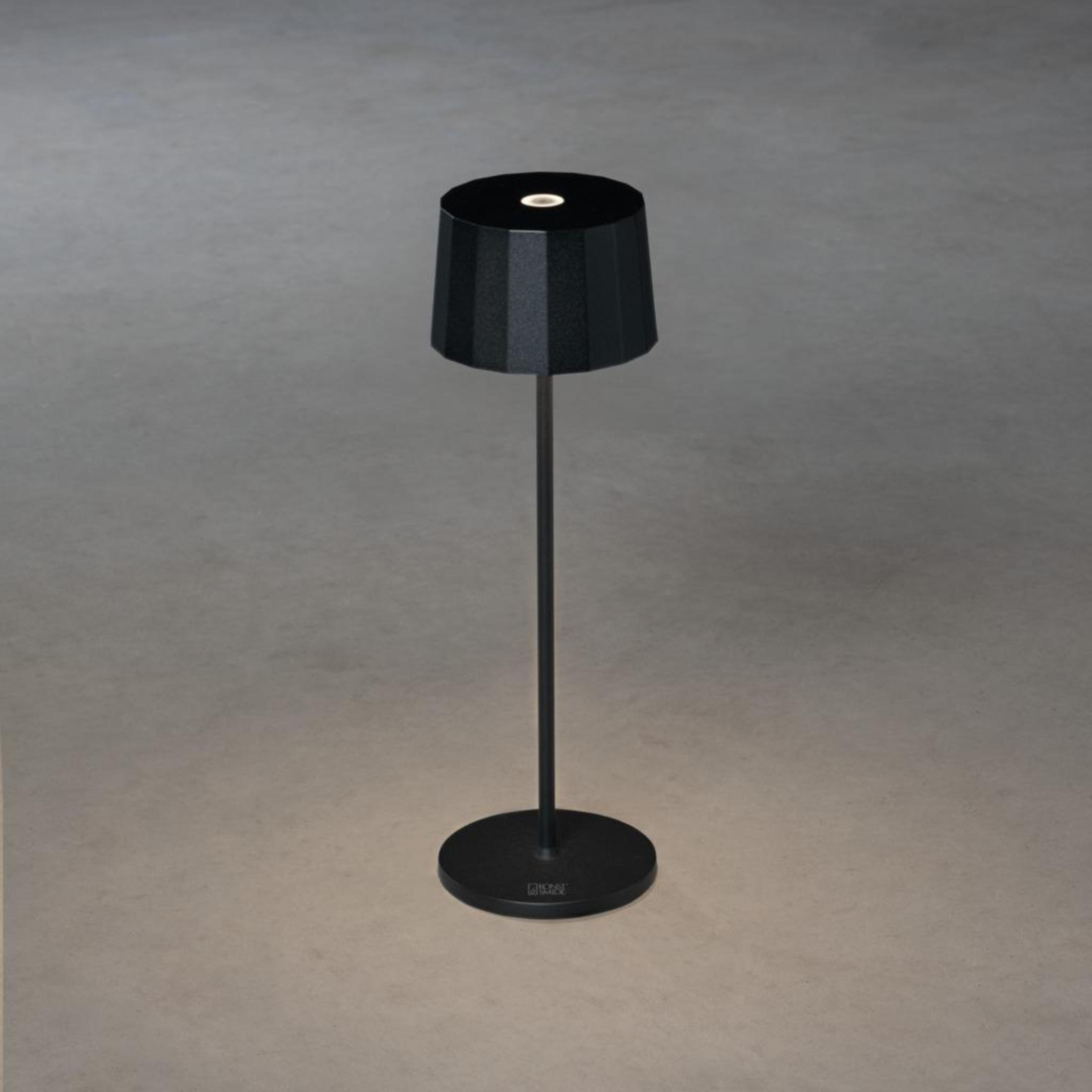 Eine stehende schwarze Lampe mit zylindrischem Sockel und einem abgestuften Lampenschirm steht auf einer neutralgrauen Oberfläche.