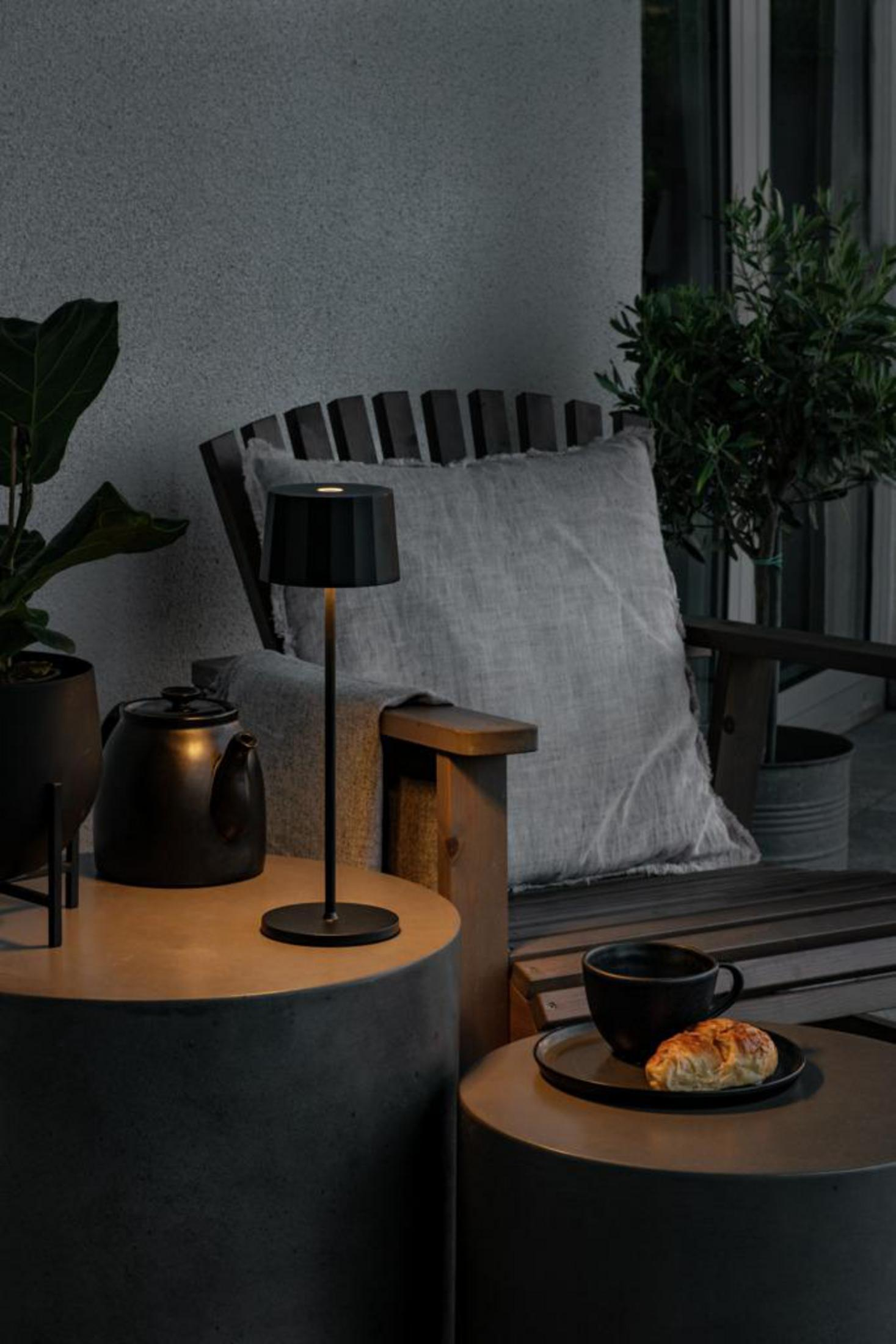 Eine schwarze Lampe steht auf einem kleinen Tisch neben einem Stuhl, mit einer Kaffeetasse, einem Croissant und einer Teekanne in einer gemütlichen Outdoor-Umgebung.