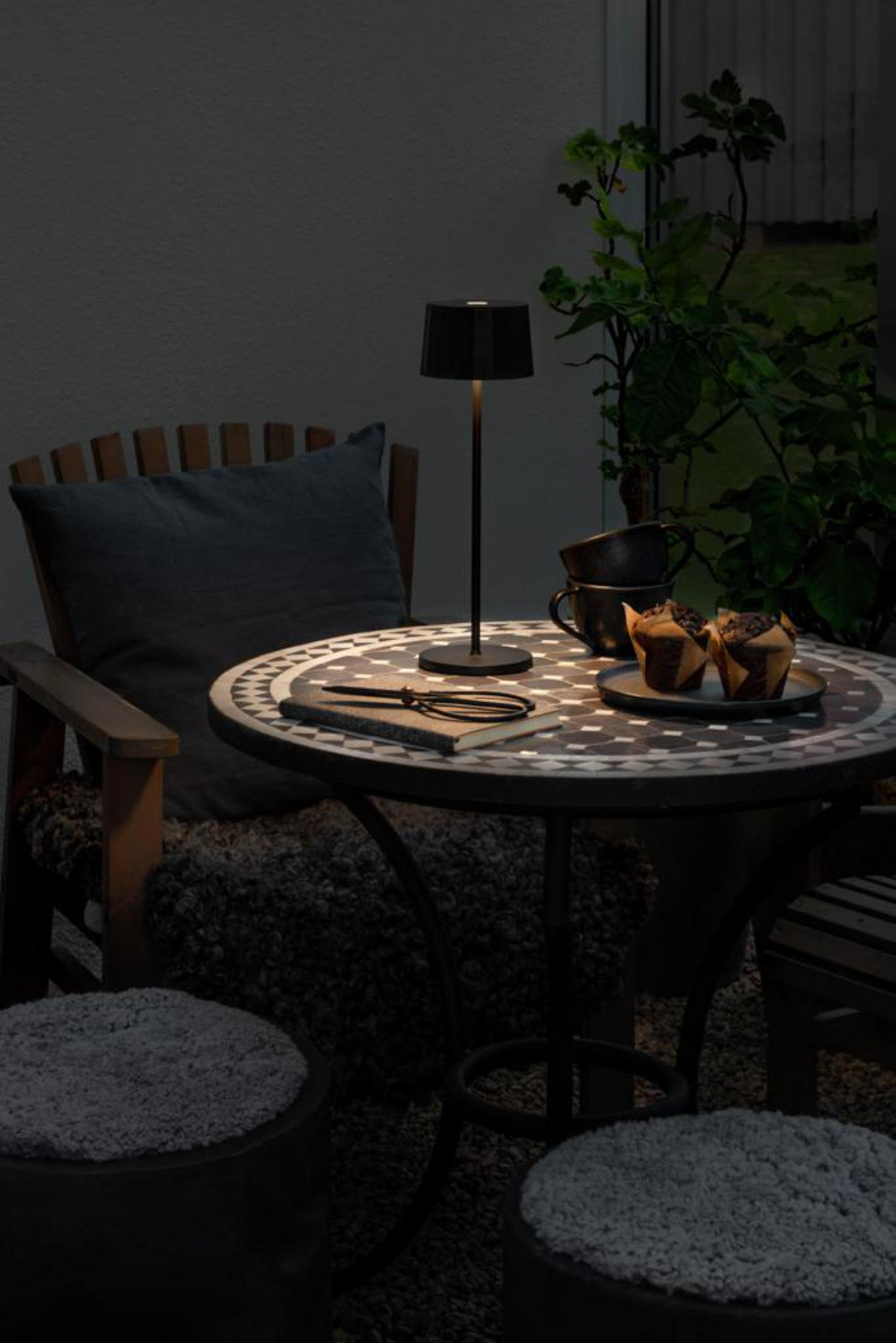 Eine schwarze Lampe auf einem Tisch mit einem Notizbuch, Muffins und Pflanzen schafft eine gemütliche Outdoor-Szene mit Stühlen und Hockern.
