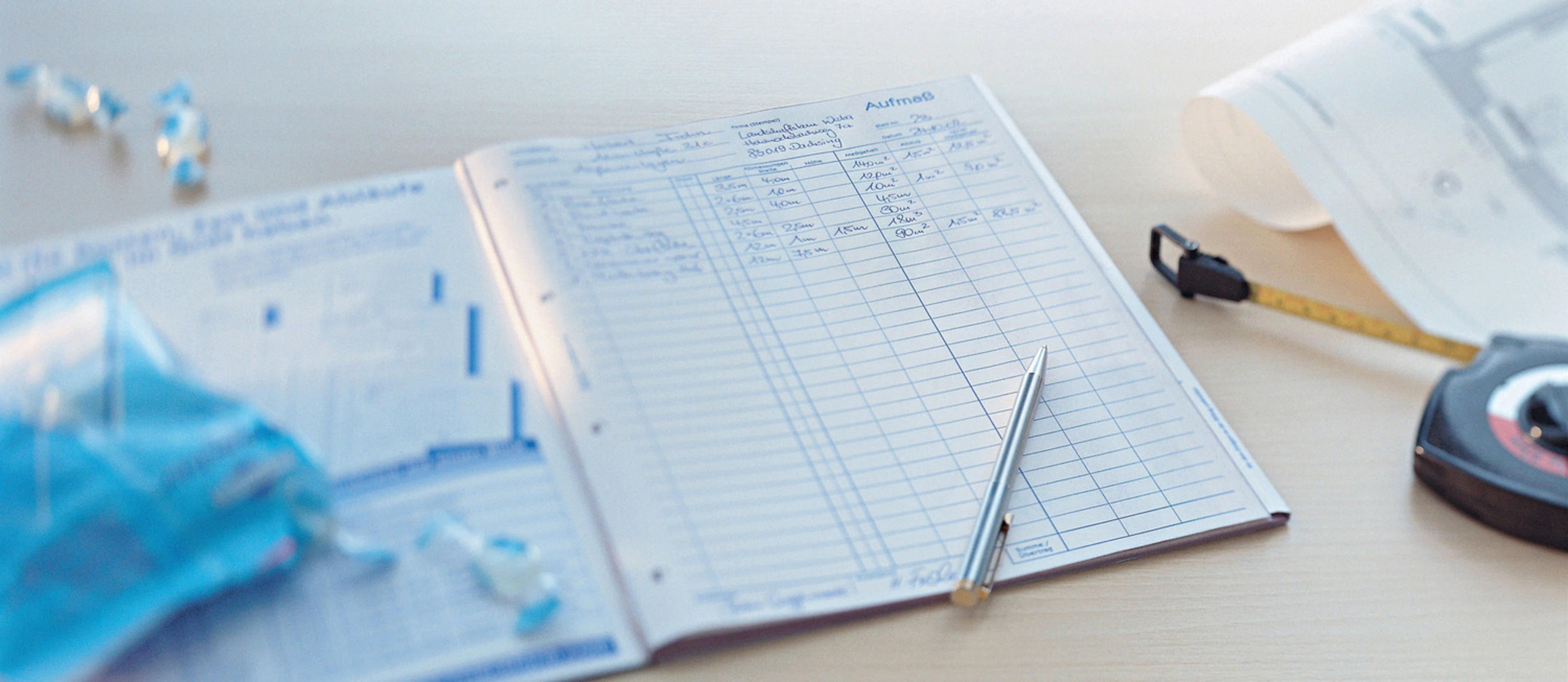 Offenes Notizbuch mit linierten Seiten, Stift, Maßband und Papierrolle auf einer hellen Holzoberfläche.