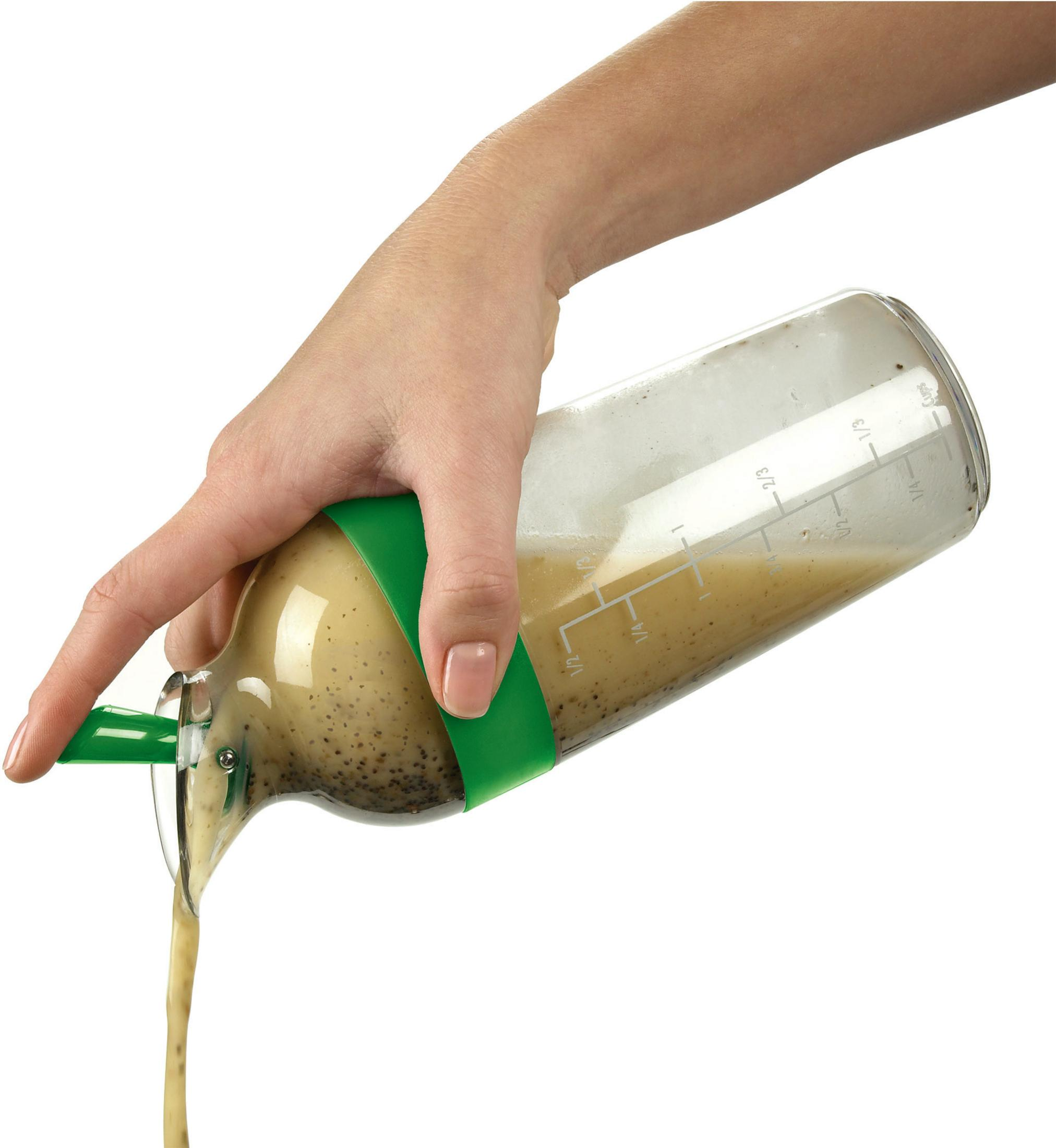 Mano che versa la vinaigrette da una bottiglia di vetro trasparente con dettagli verdi, su uno sfondo bianco brillante. Marcature di misurazione visibili.