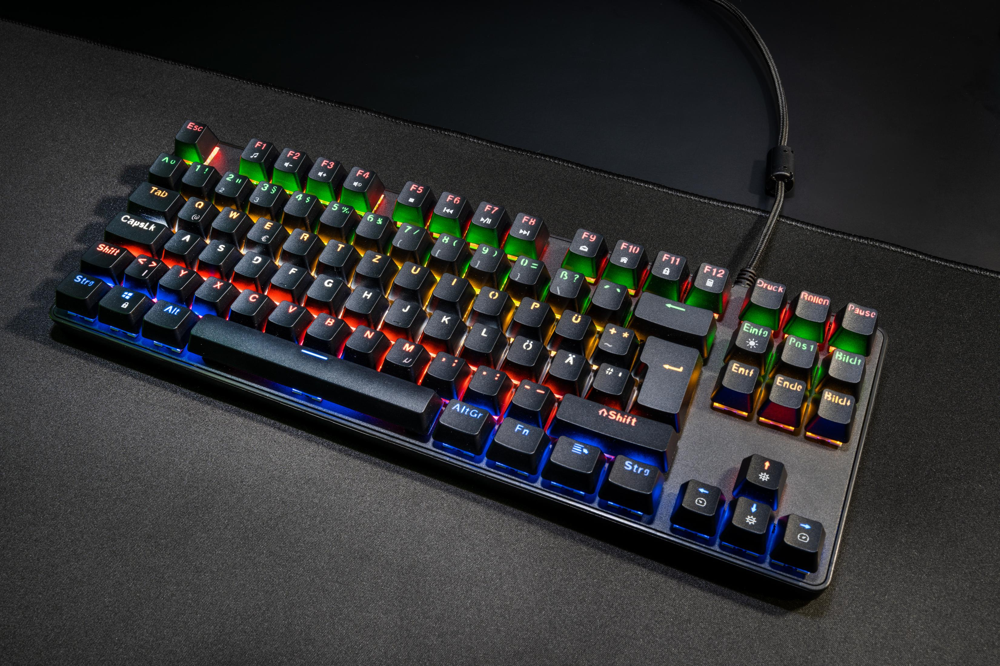 Schwarze Tastatur mit beleuchteten Tasten, schräg auf dunkler Oberfläche. Tastenfarben sind rot, orange, gelb, grün, blau.