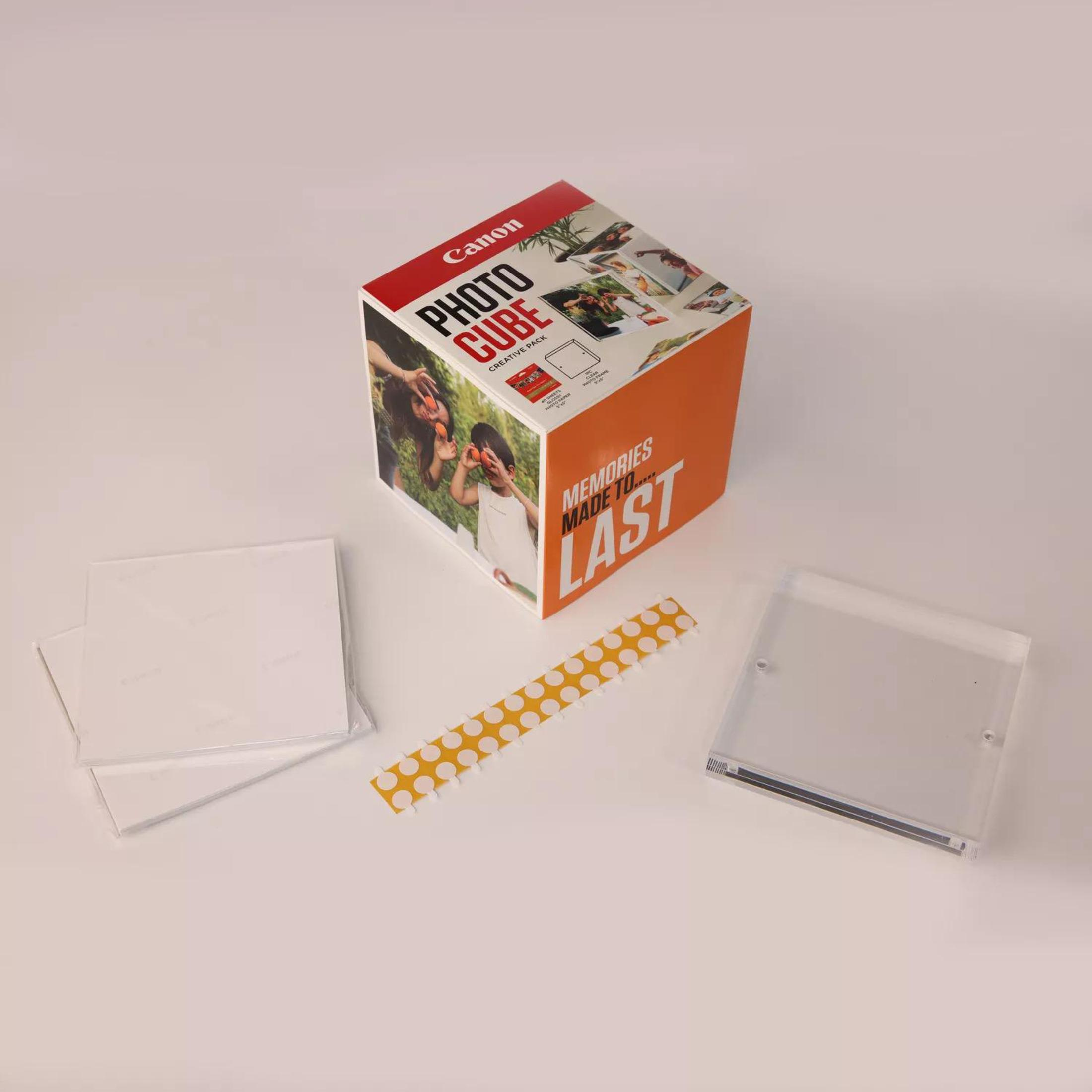 Una scatola Canon Photo Cube con foto, cornici trasparenti e una striscia di adesivi, su una superficie bianca.