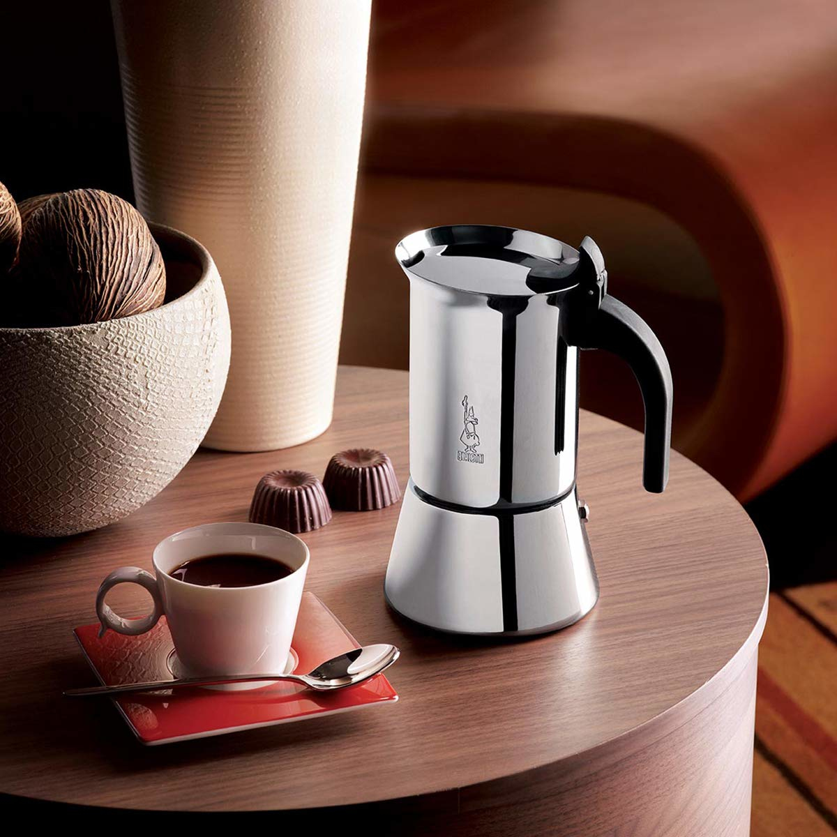 Una moka in acciaio inossidabile su un tavolo con una tazza di caffè e cioccolatini.