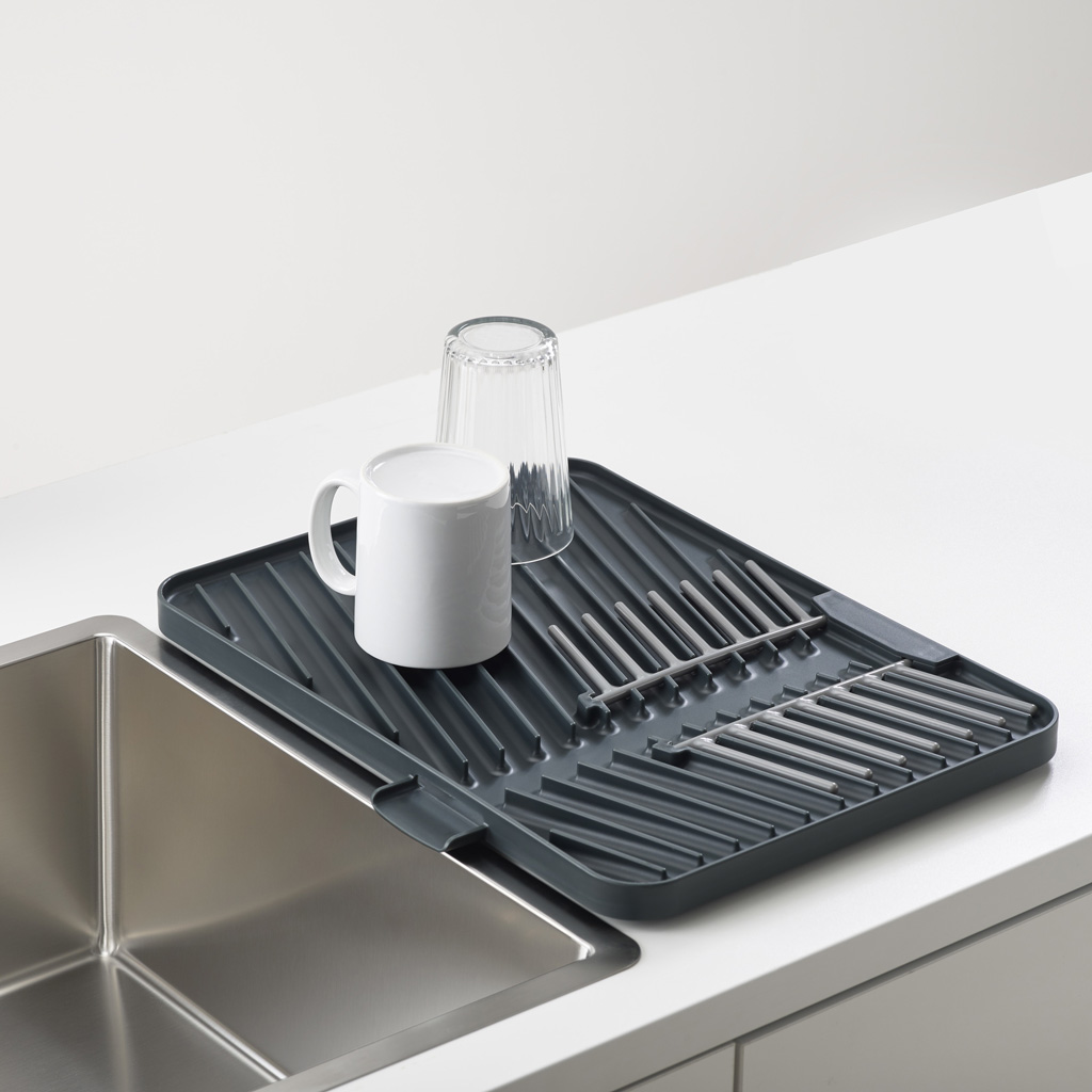 Un escurridor gris junto a un fregadero sostiene una taza blanca y un vaso transparente sobre una encimera blanca.