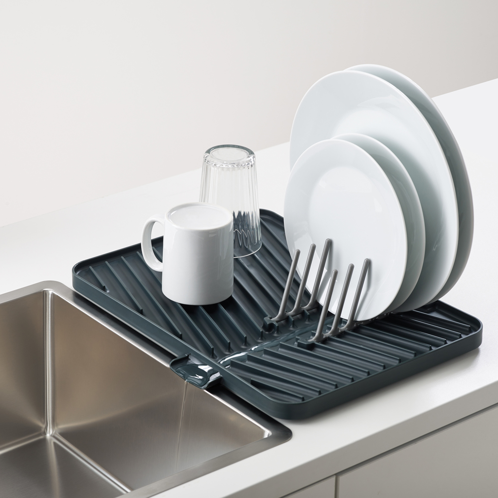 Un escurridor gris sostiene platos, un vaso y una taza junto a un fregadero de acero inoxidable sobre una encimera blanca.