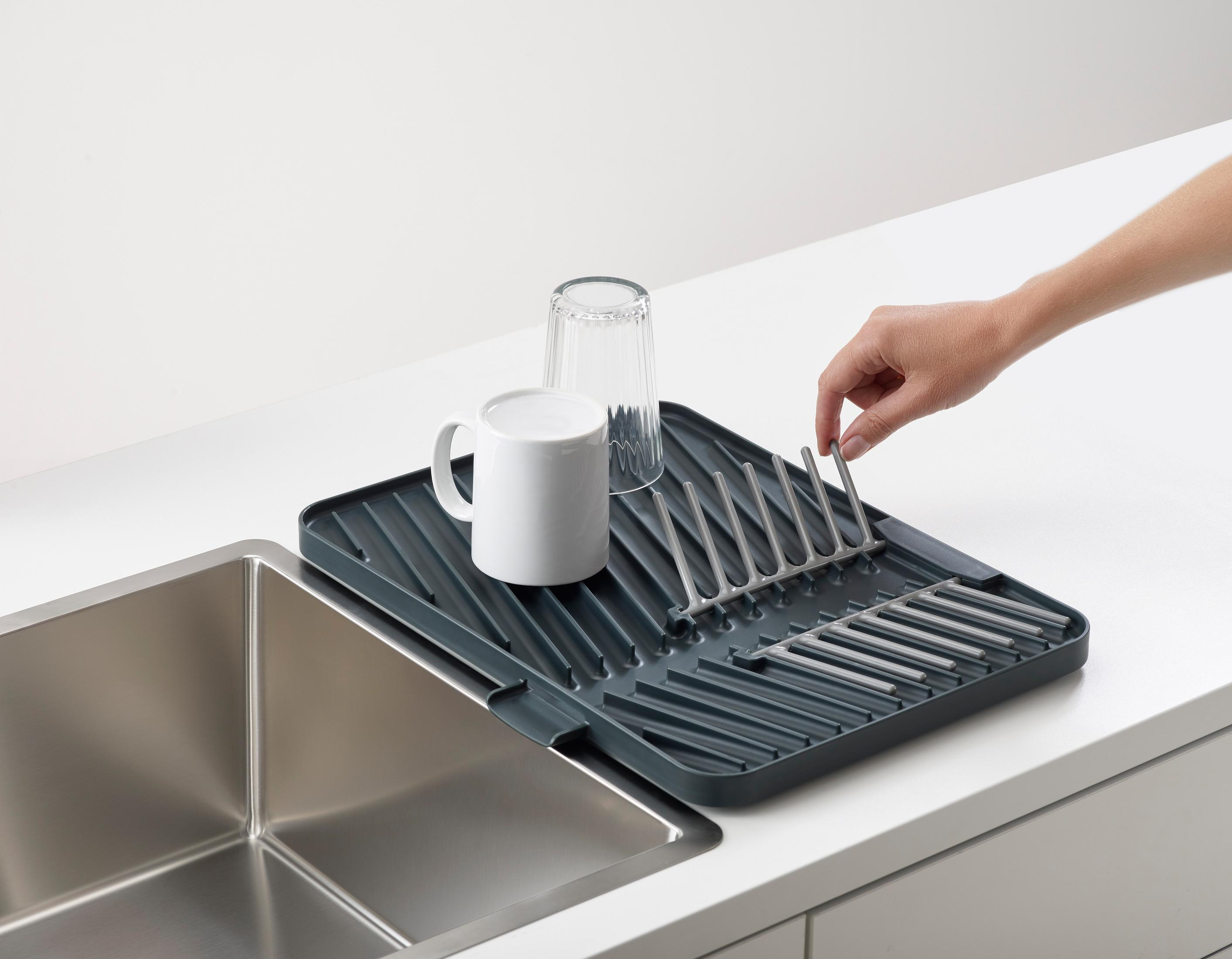 Un escurridor gris con una taza blanca y un vaso junto a un fregadero de acero inoxidable, con una mano ajustando un estante.