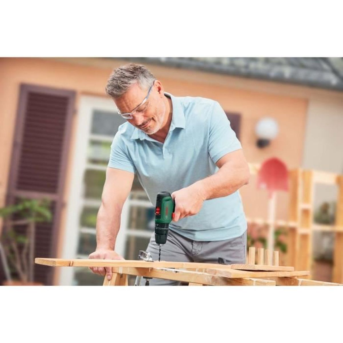 Un uomo fora il legno all'aperto con un trapano Bosch verde, indossando occhiali di sicurezza, con una tettoia sullo sfondo.