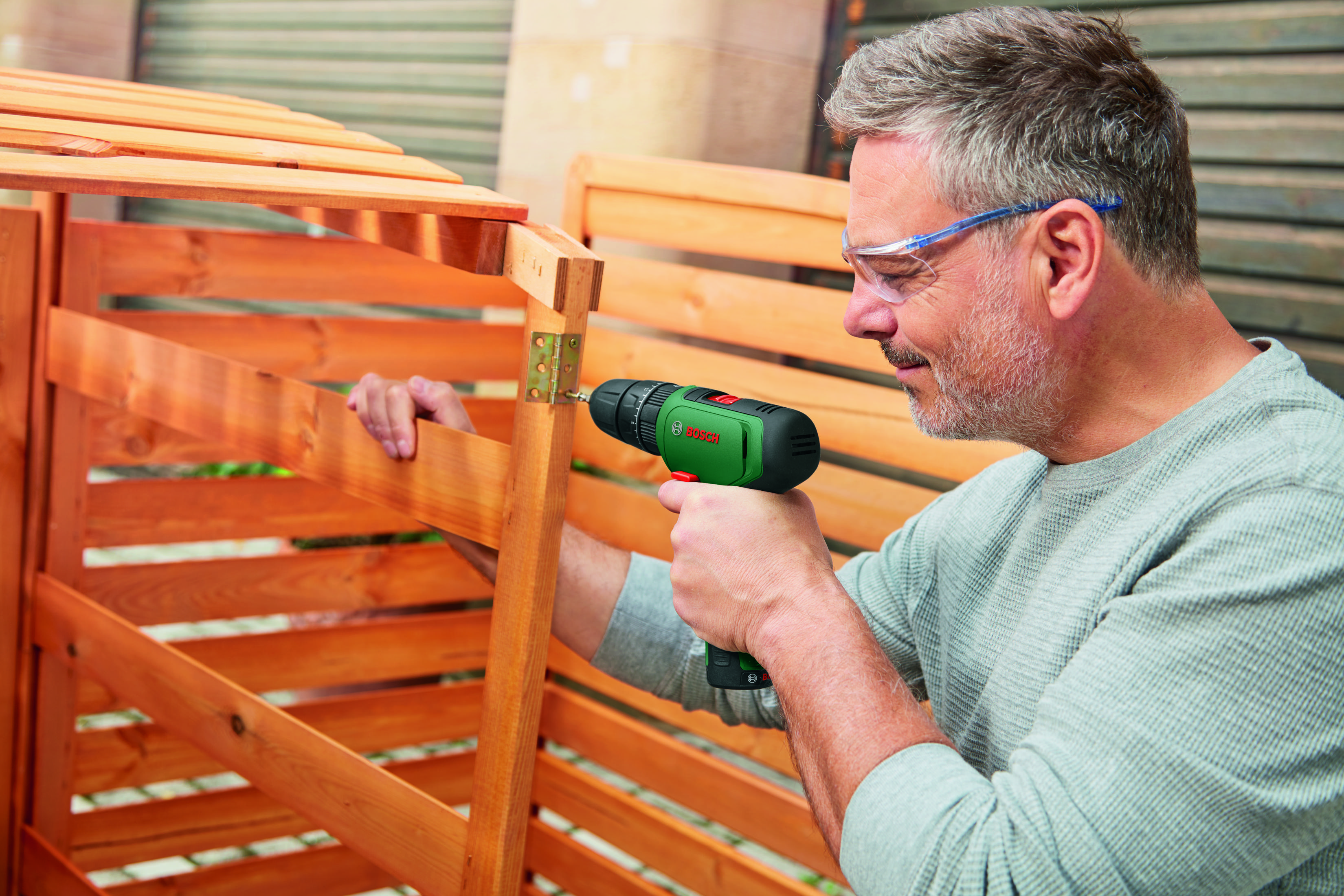 Un uomo che indossa occhiali di sicurezza fora una struttura in legno con un trapano Bosch verde.