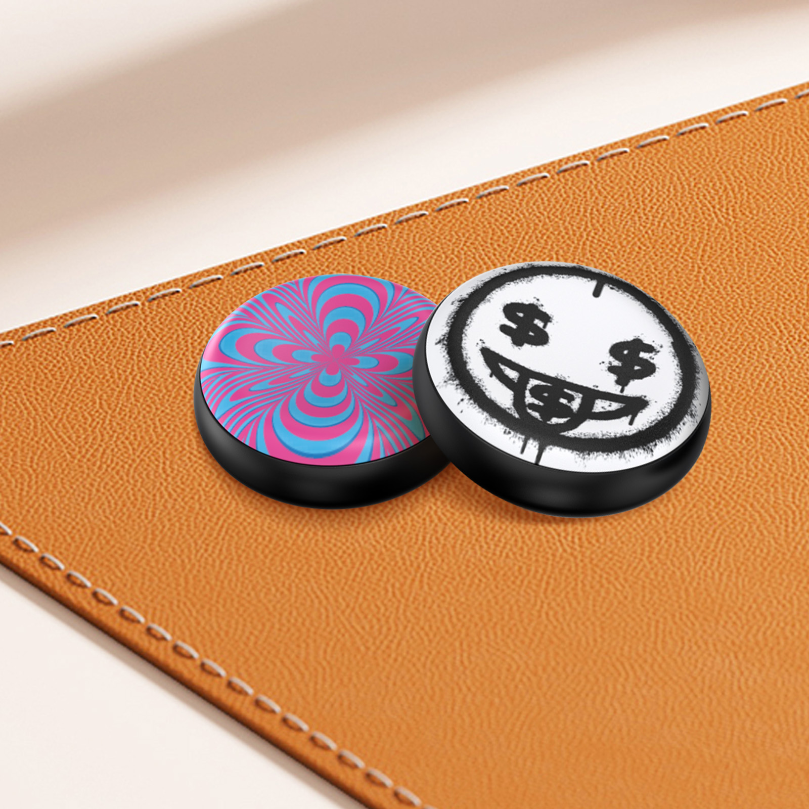 Due PopSocket magnetici rotondi, uno con un vortice e l'altro con una faccina sorridente su una superficie marrone.