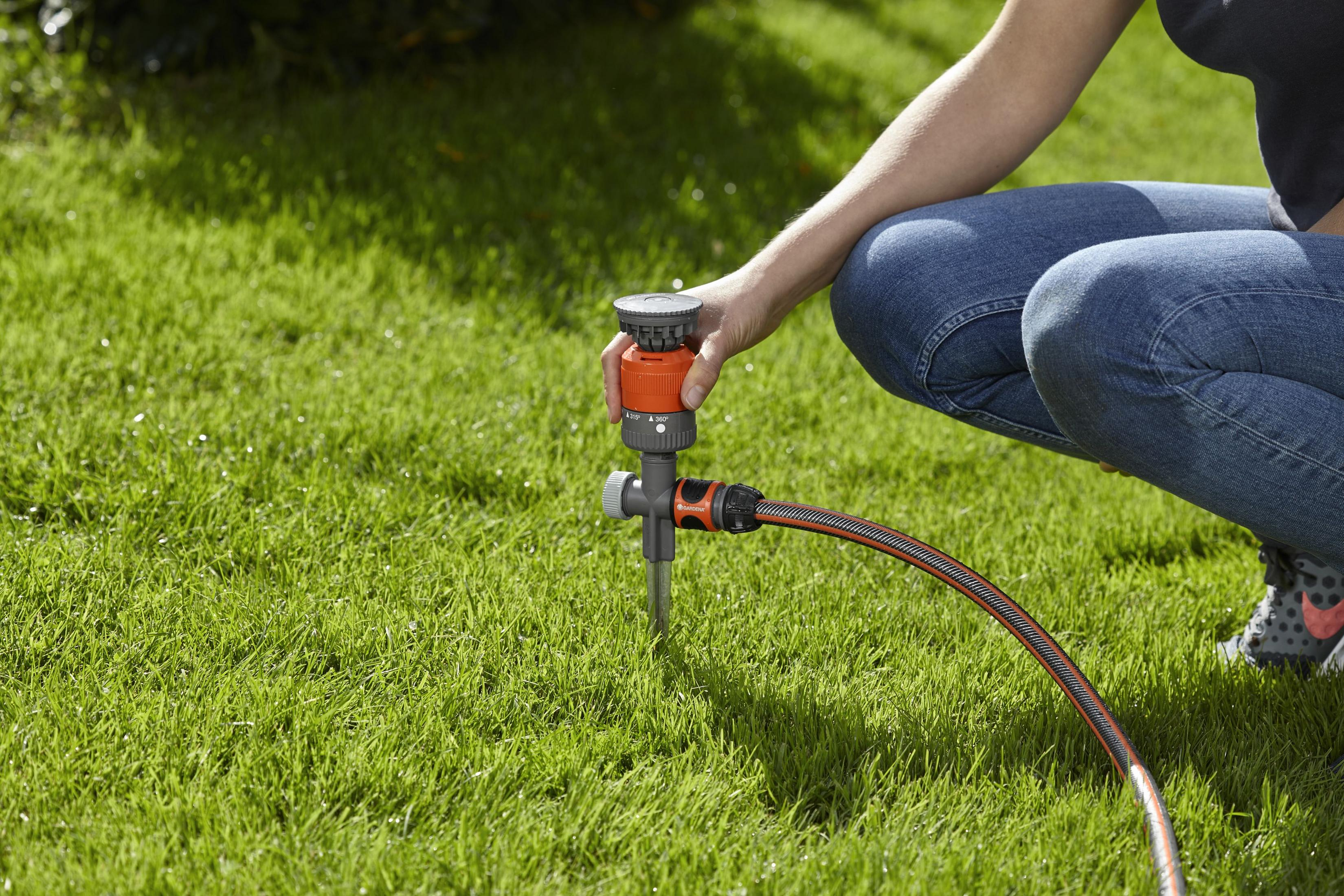 Una donna si inginocchia per regolare un irrigatore grigio e arancione attaccato a un tubo sull'erba.