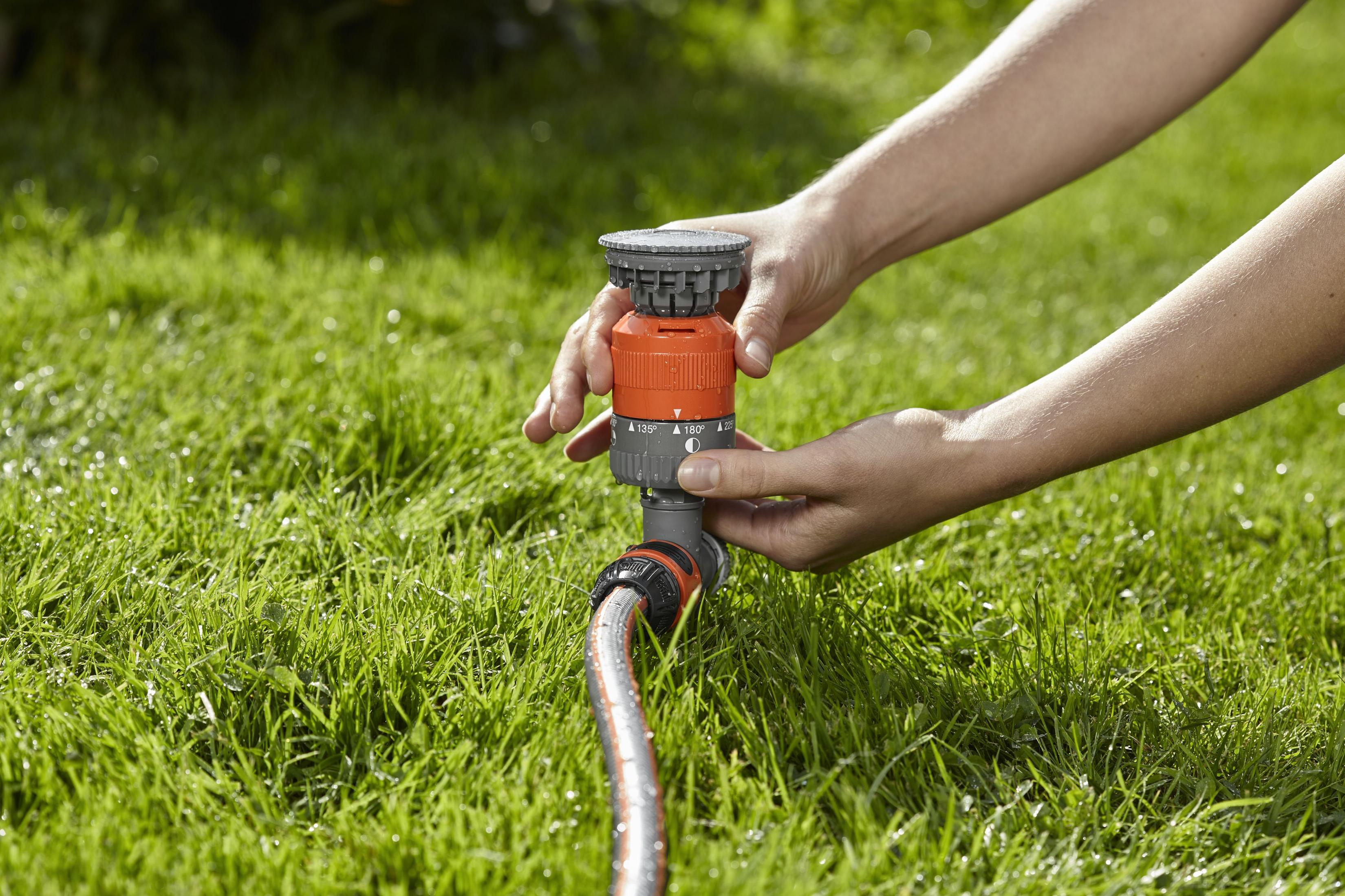 Le mani regolano un attacco per tubo da giardino arancione e grigio su erba verde, mostrando gocce d'acqua.
