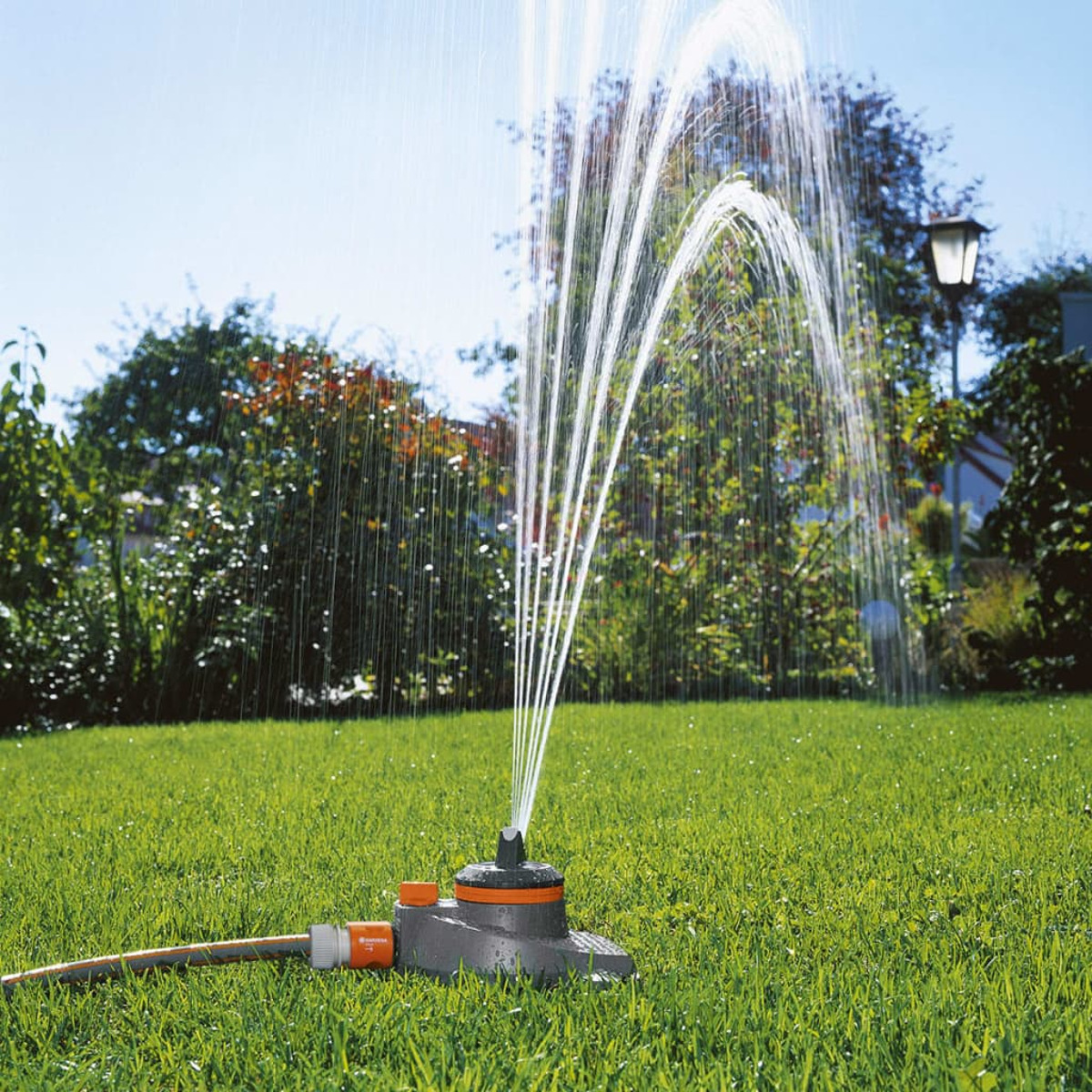 Ein Rasensprenger spritzt Wasser auf grünes Gras unter blauem Himmel. Im Hintergrund ist ein Garten.