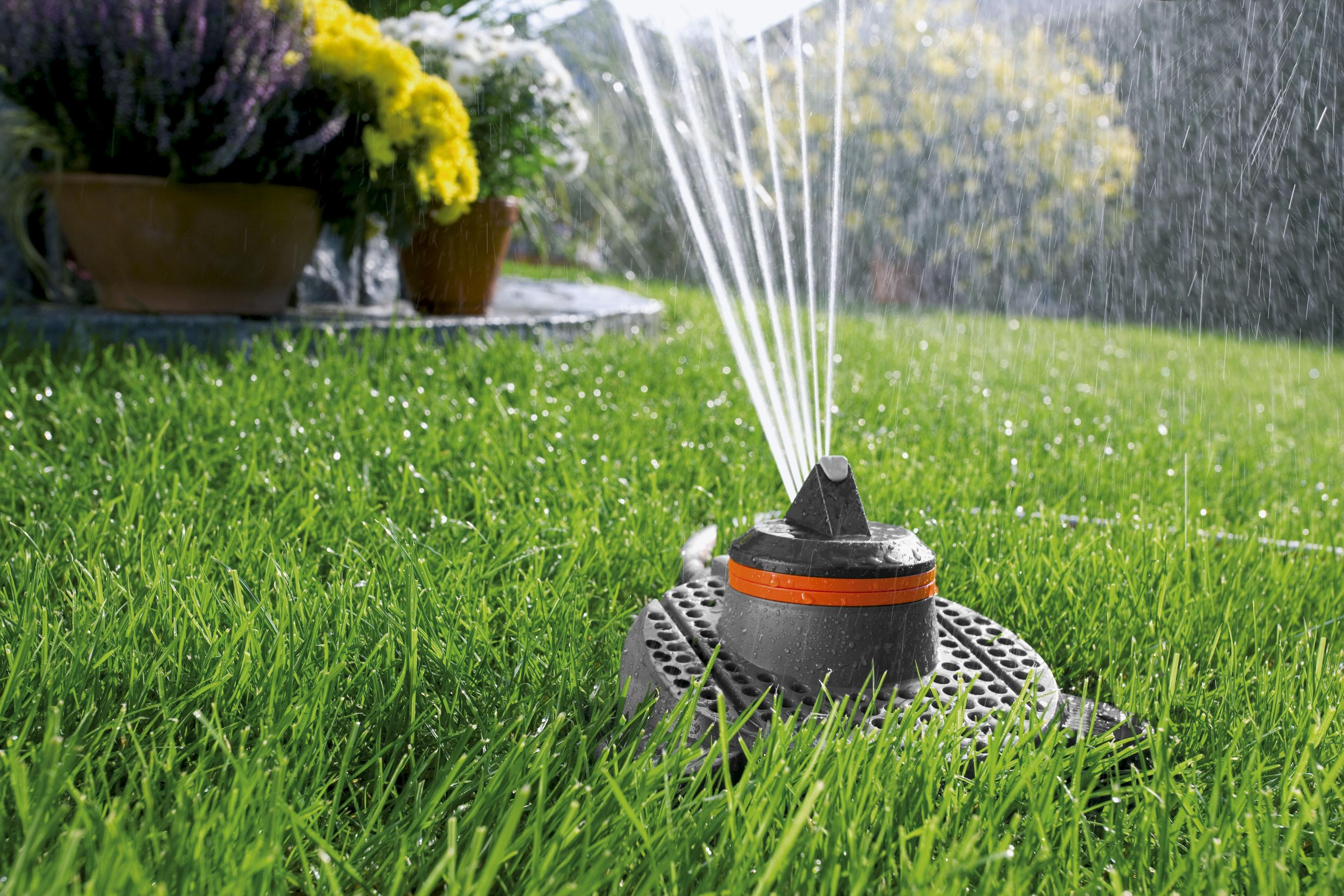 Ein Rasensprenger versprüht Wasser über grünes Gras. Blumen und Pflanzen sind im Hintergrund.