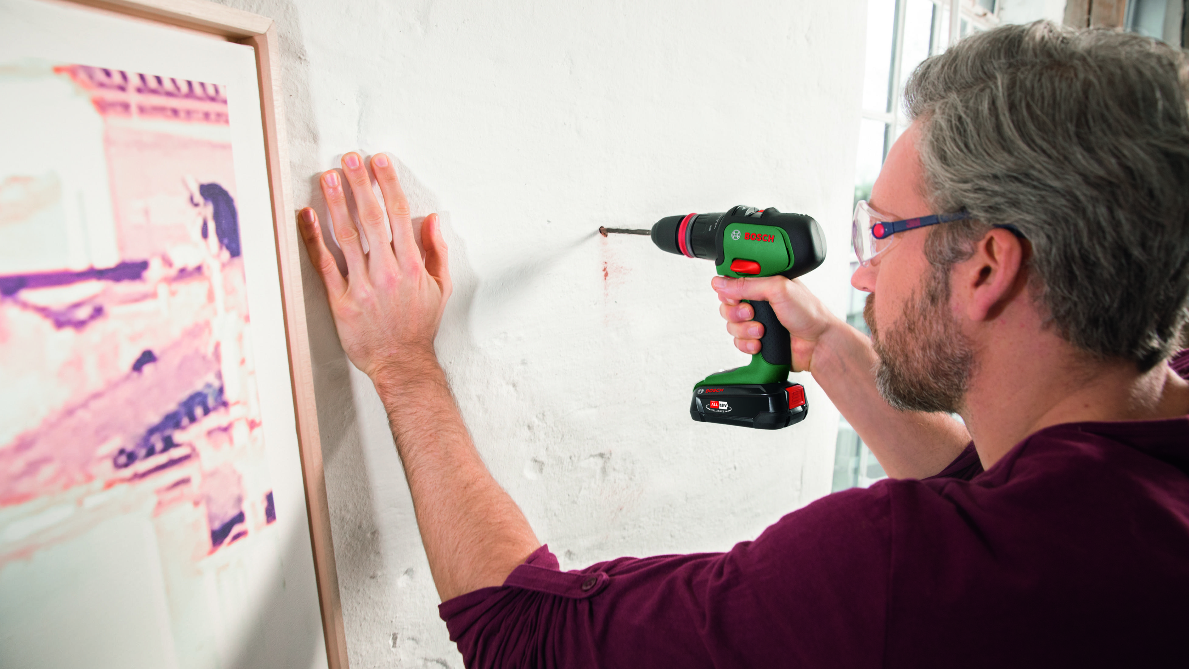 Un uomo fora un muro con un trapano avvitatore Bosch. Indossa occhiali e un maglione bordeaux.