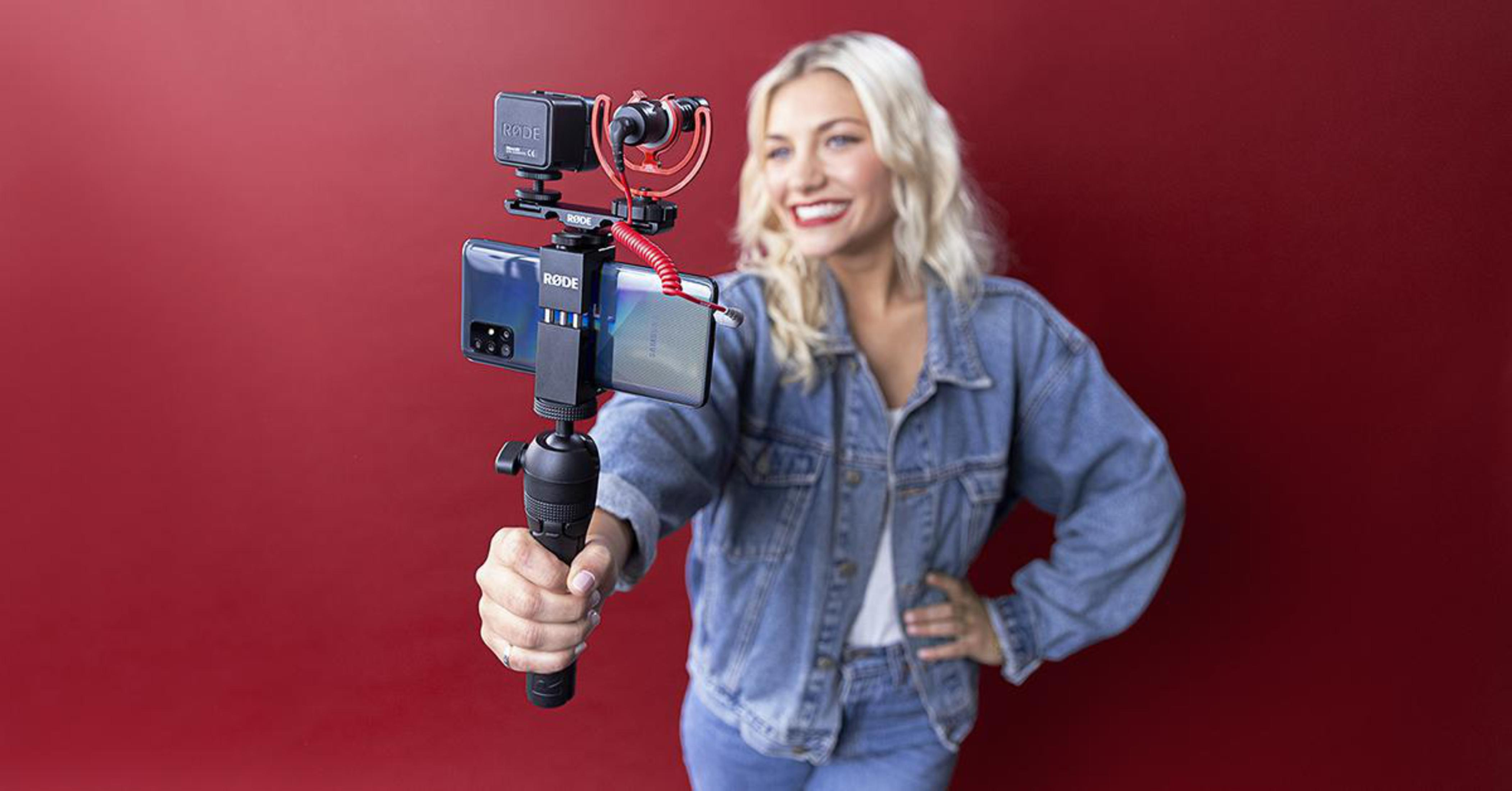 Donna con telefono e microfono, sfondo rosso. Capelli biondi, sorridente, giacca di jeans.