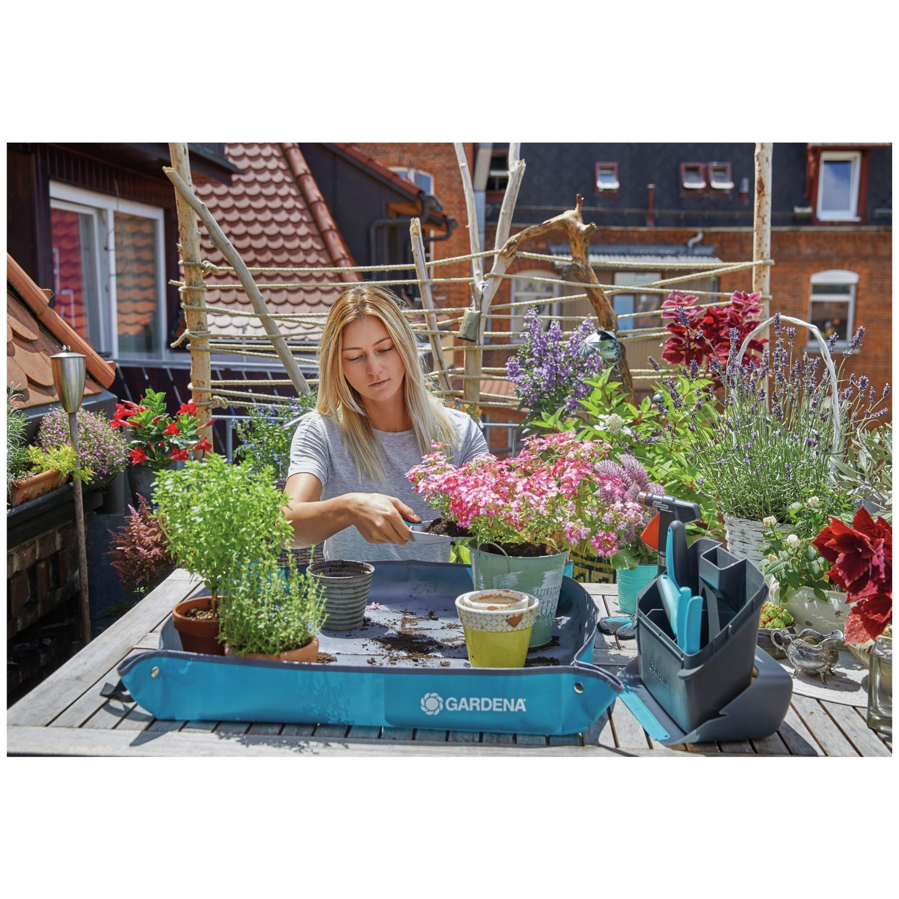 Frau gärtnert auf einer blauen GARDENA-Matte, umgeben von Pflanzen, Werkzeugen und einer Dachlandschaft.