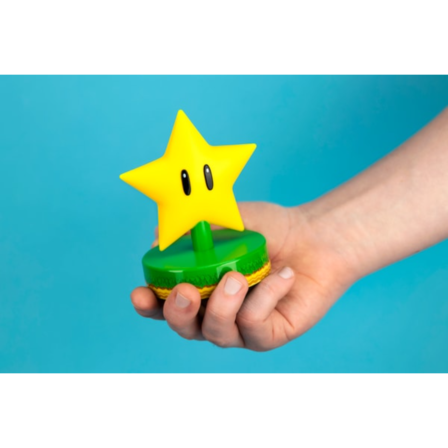 Ręka trzyma żółtą figurkę gwiazdy Super Mario z czarnymi oczami na zielonej podstawie.