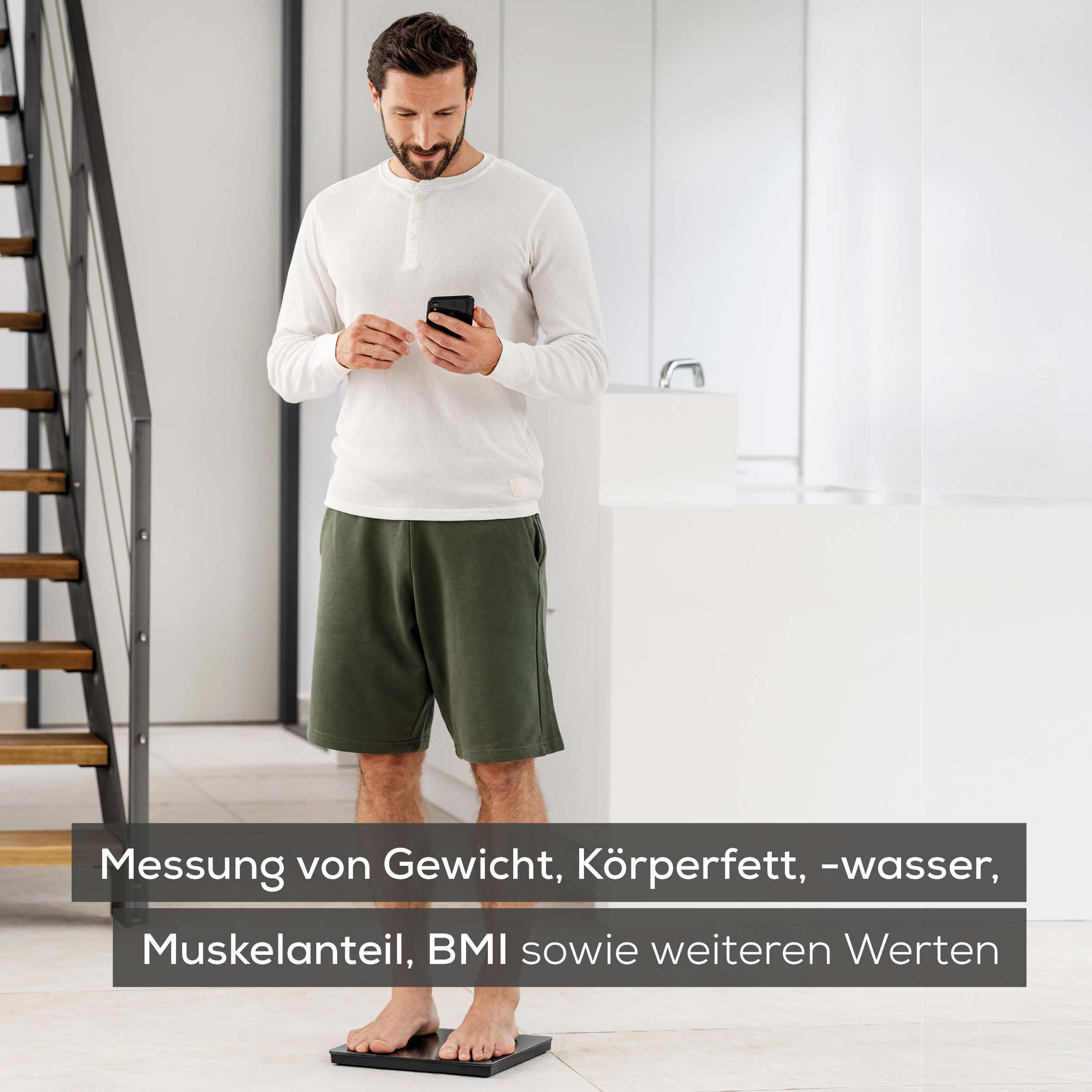 Ein Mann steht auf einer Waage und hält ein Telefon. Die Waage ist schwarz. Küche und Treppe im Hintergrund.