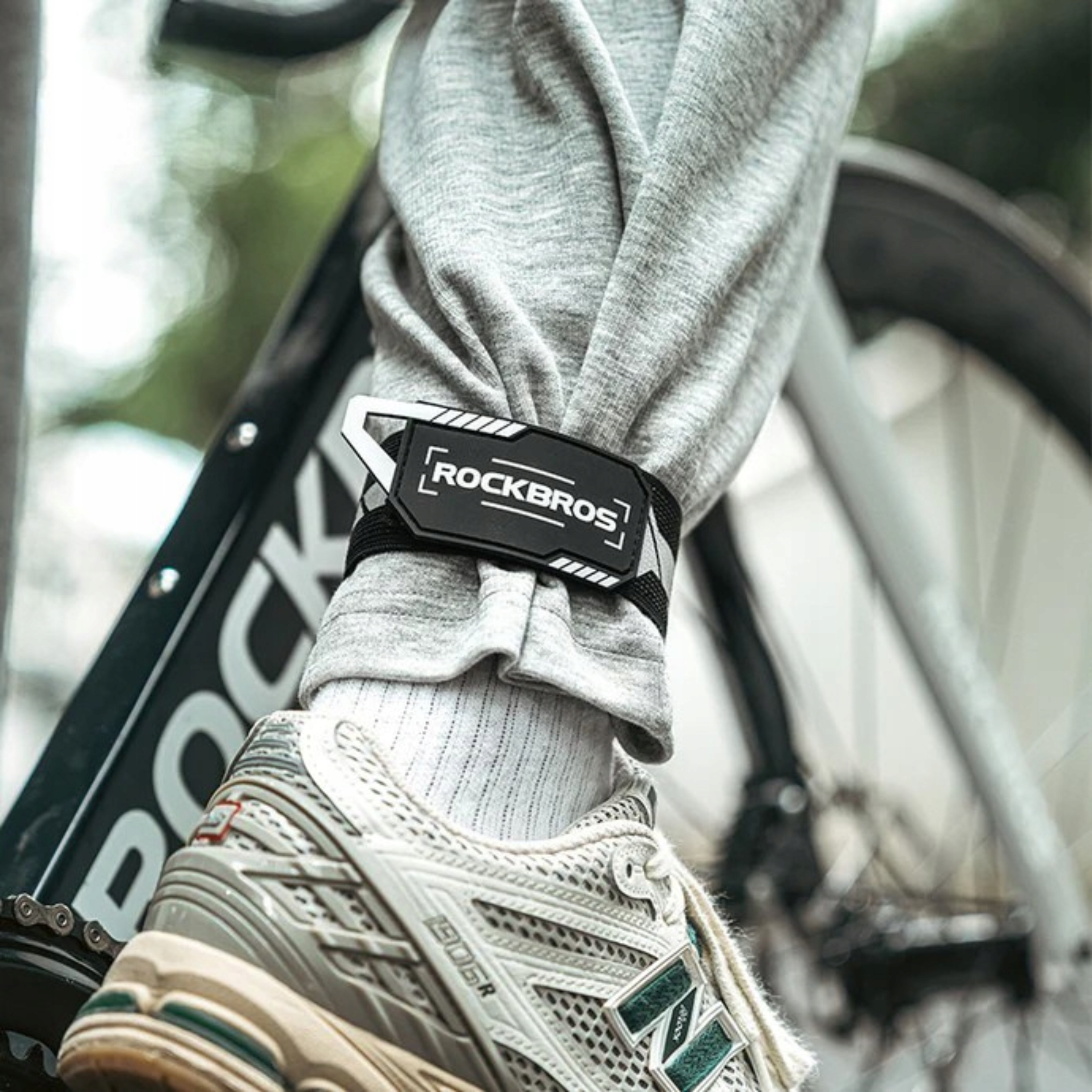 Noga z opaską Rockbros wokół kostki w pobliżu roweru. Osoba ma na sobie szare spodnie i trampki.