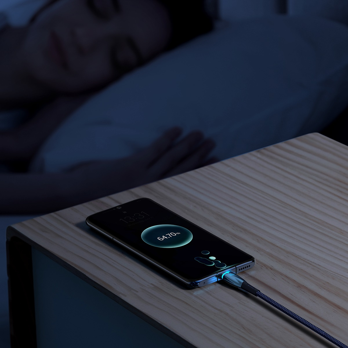 Telefon ładujący się kablem, pokazujący 64.70% i śpiąca osoba w tle.