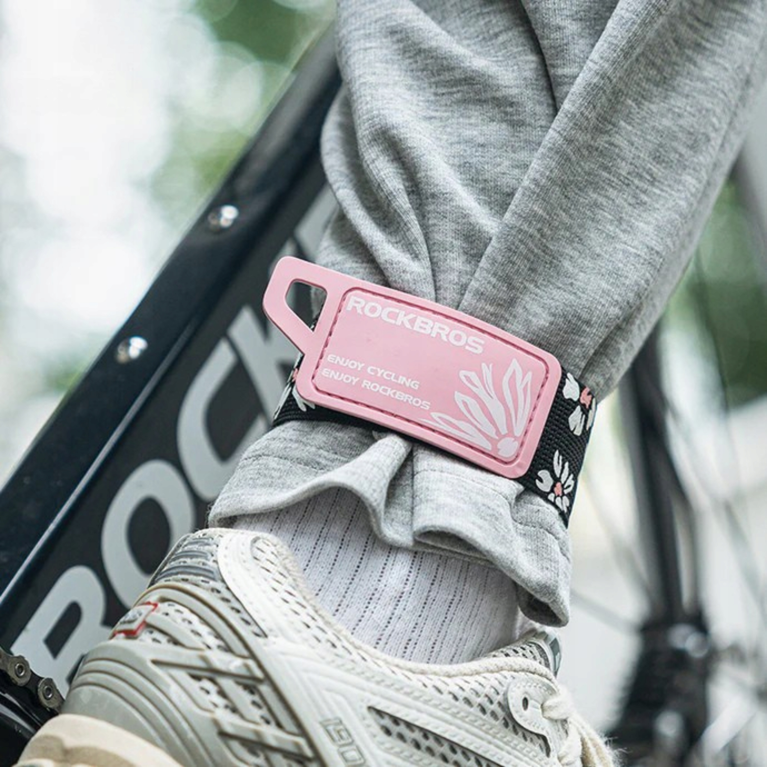 Noga osoby z różową opaską na nadgarstek ROCKBROS, wokół kostki, wraz z rowerem w tle.