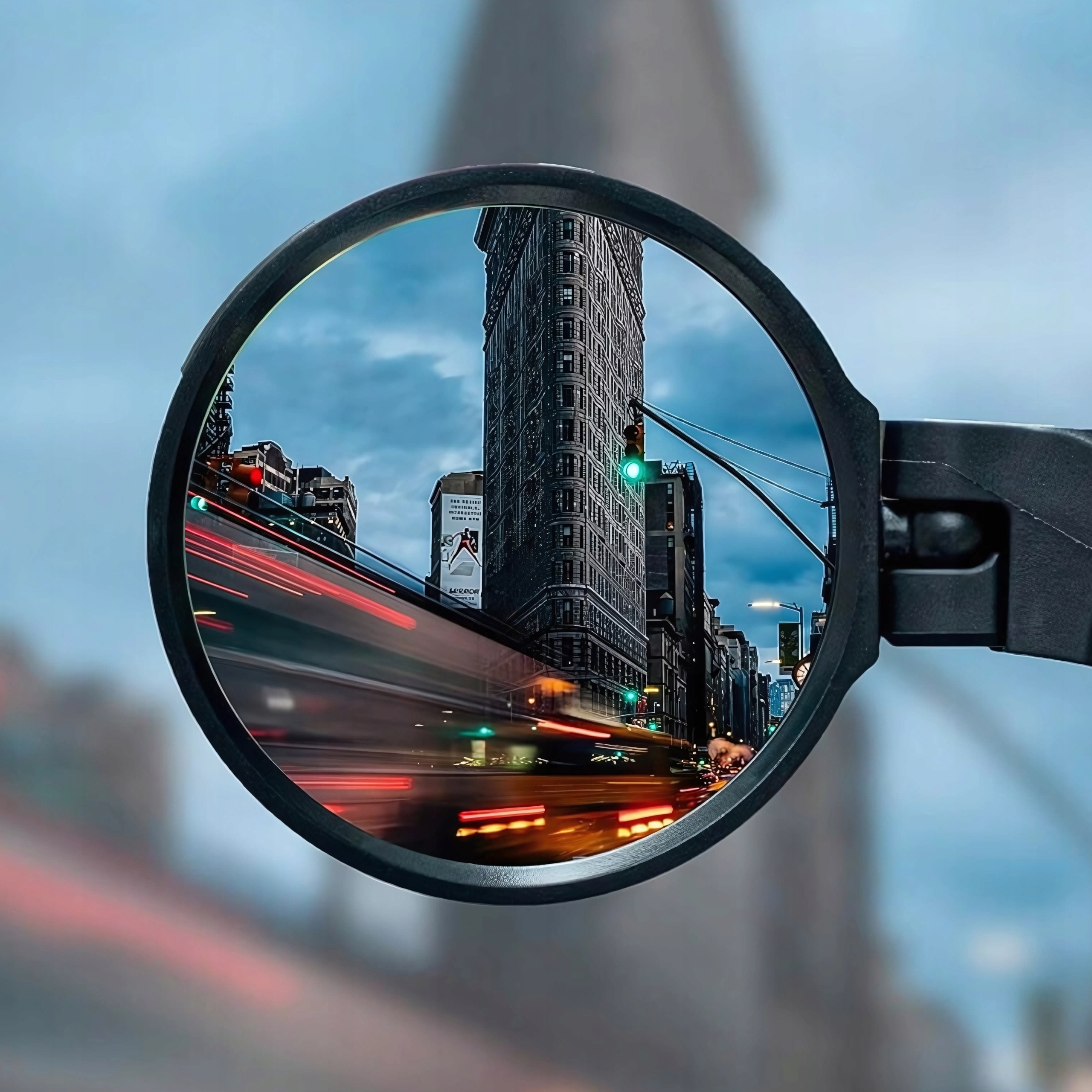 Lusterko rowerowe odbijające scenę uliczną miasta z wysokim budynkiem i rozmytymi światłami.